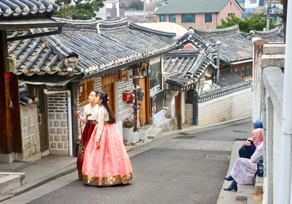 Two women in traditional hanbok dresses walking on a narrow street in a Korean neighborhood with tiled rooftops and sitting women in modern clothing on the side