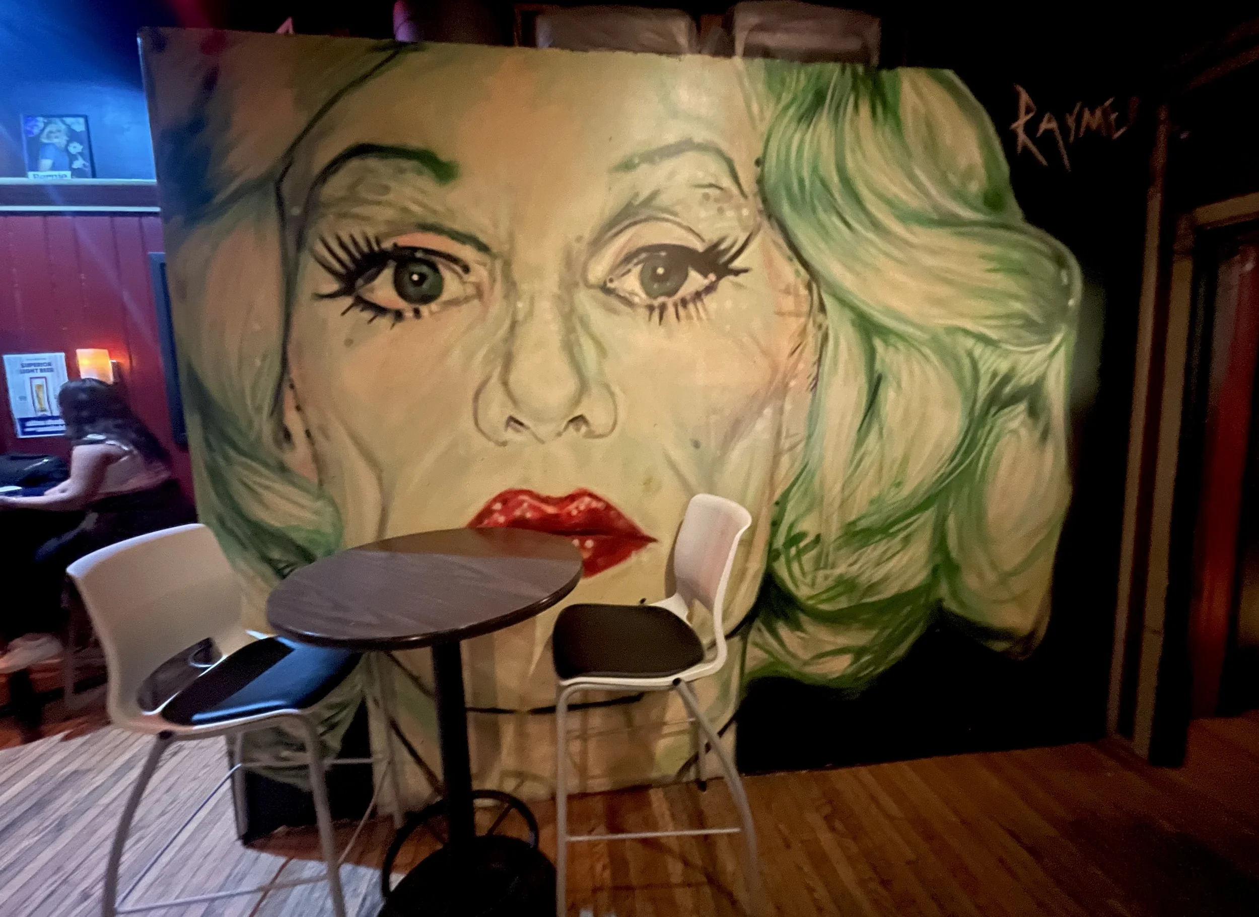 A large mural of a woman's face with green hair, red lips, and detailed eyes, painted on a wall in a dimly lit establishment with wooden floors and sitting area.