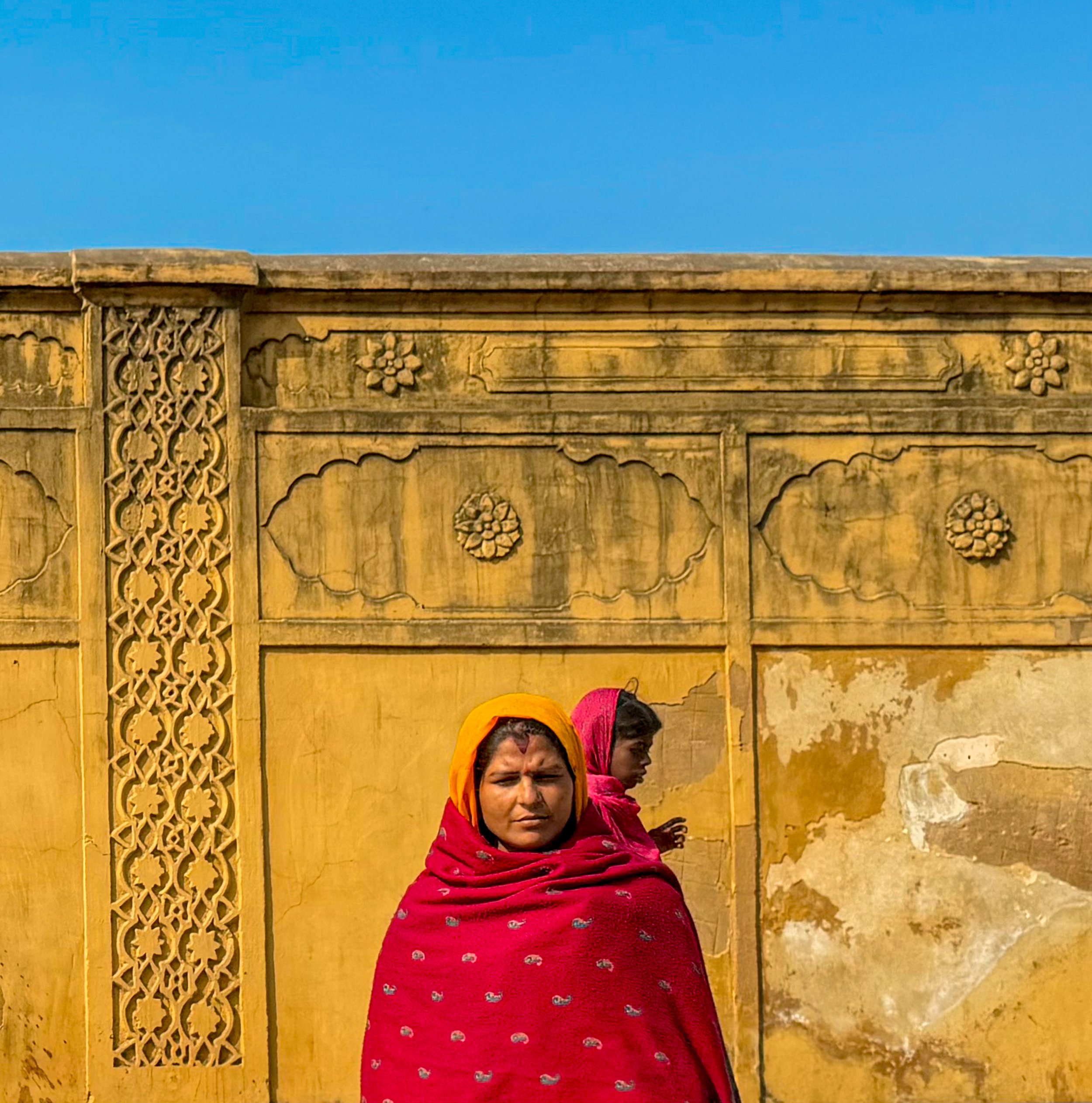 Two women in colorful sarees standing in front of a yellow weathered wall with intricate carvings and a bright blue sky above.