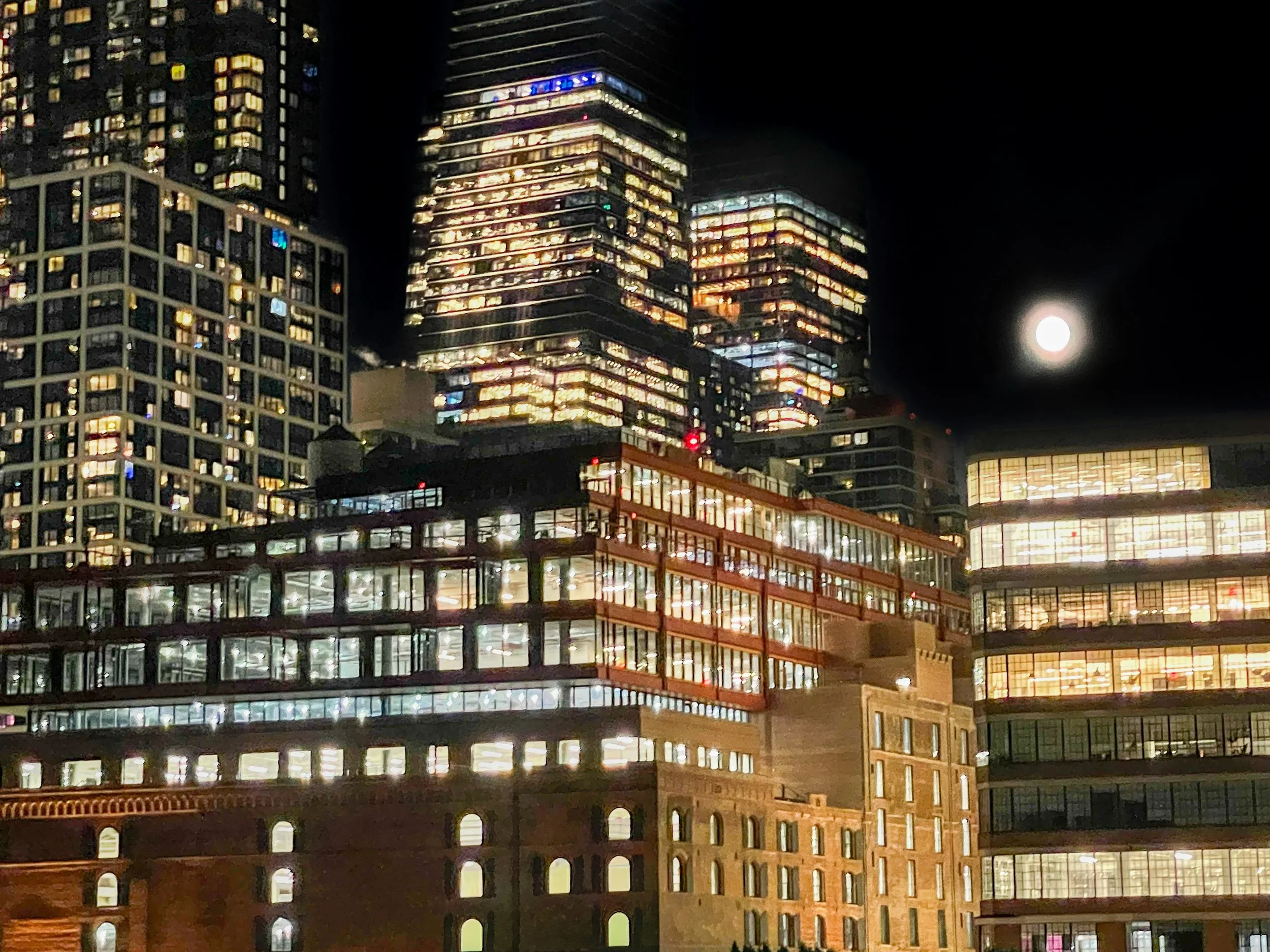Nighttime cityscape with illuminated modern glass buildings and a full moon in the sky.