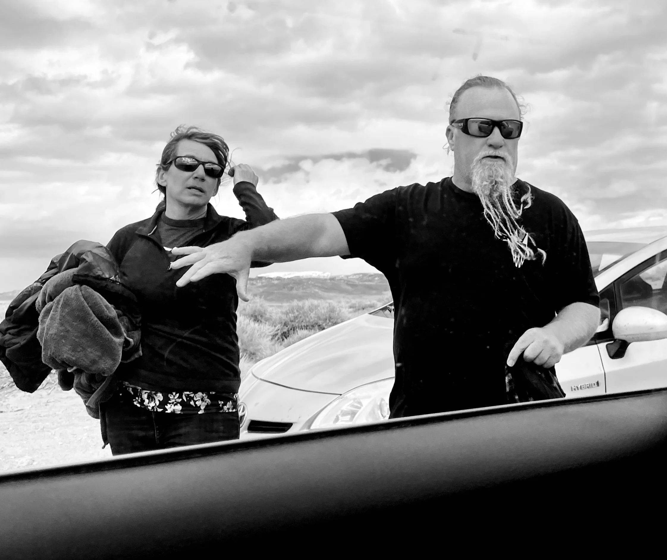 Two people wearing sunglasses standing outdoors near parked cars, with a landscape and cloudy sky in the background.
