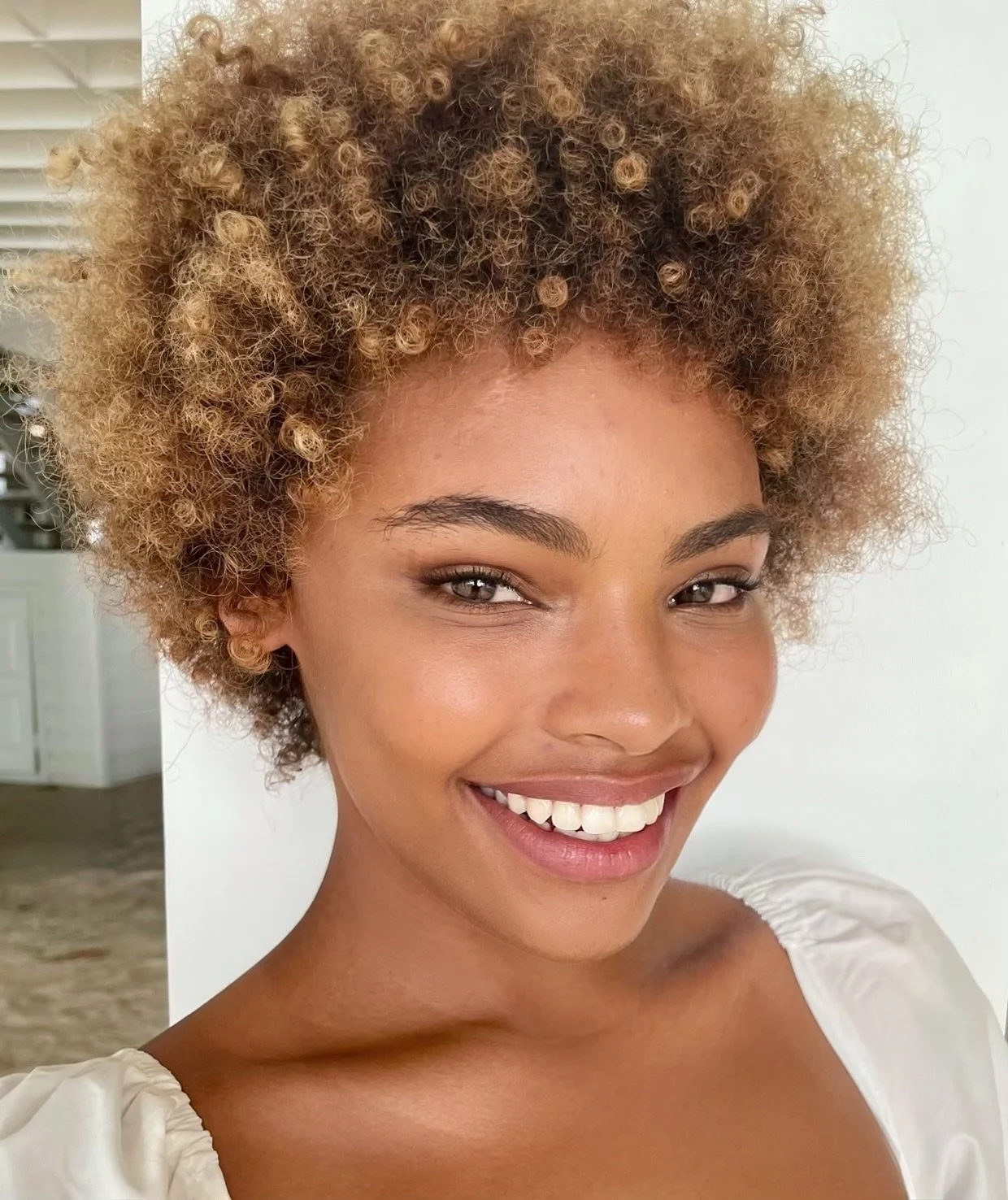 Close-up of a smiling woman with light brown skin and natural curly hair in shades of blonde and brown
