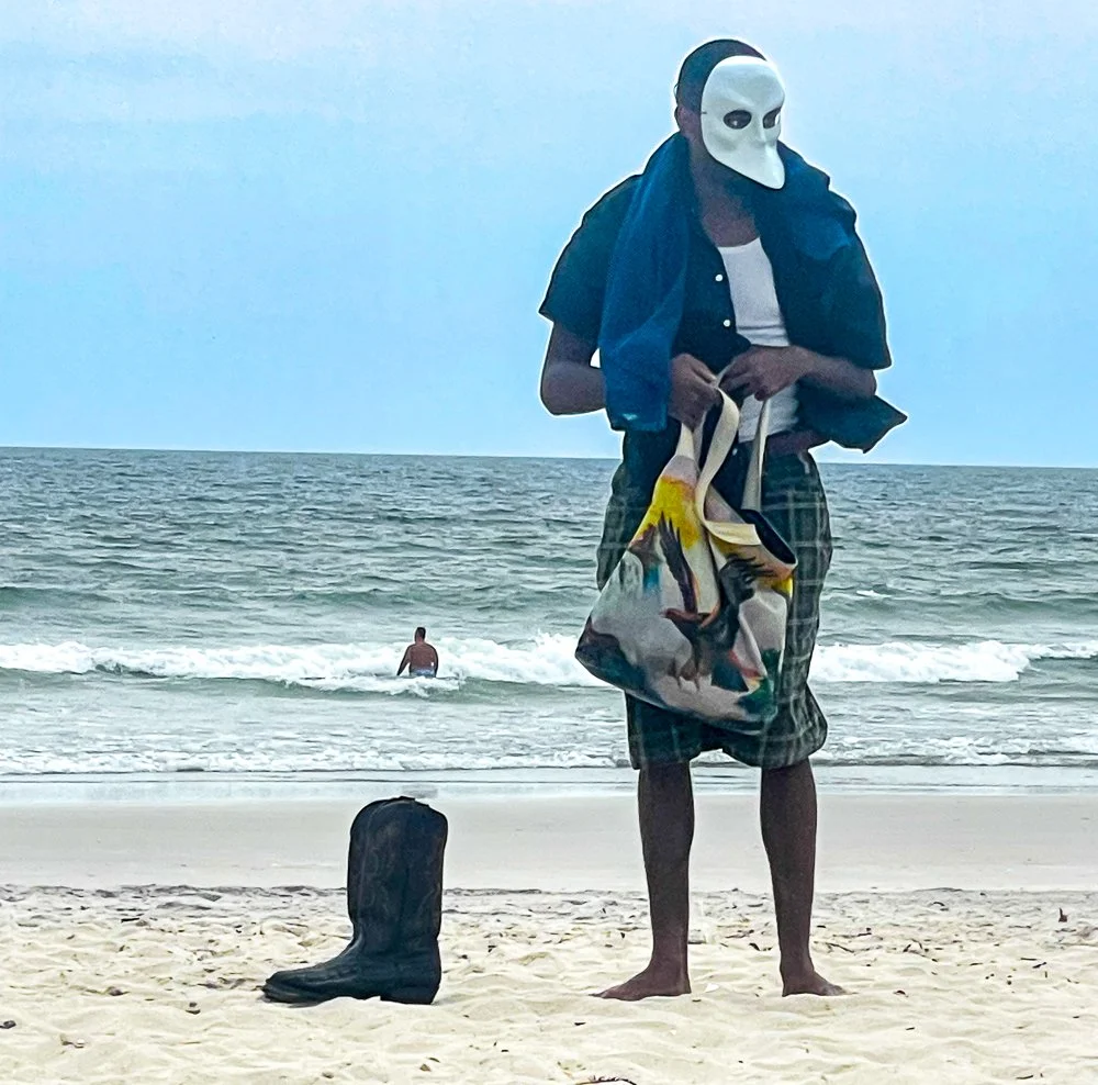 Person wearing a white mask with black eyes holding a bag and standing on a sandy beach with a boot nearby. The ocean and a person in the water are visible in the background.
