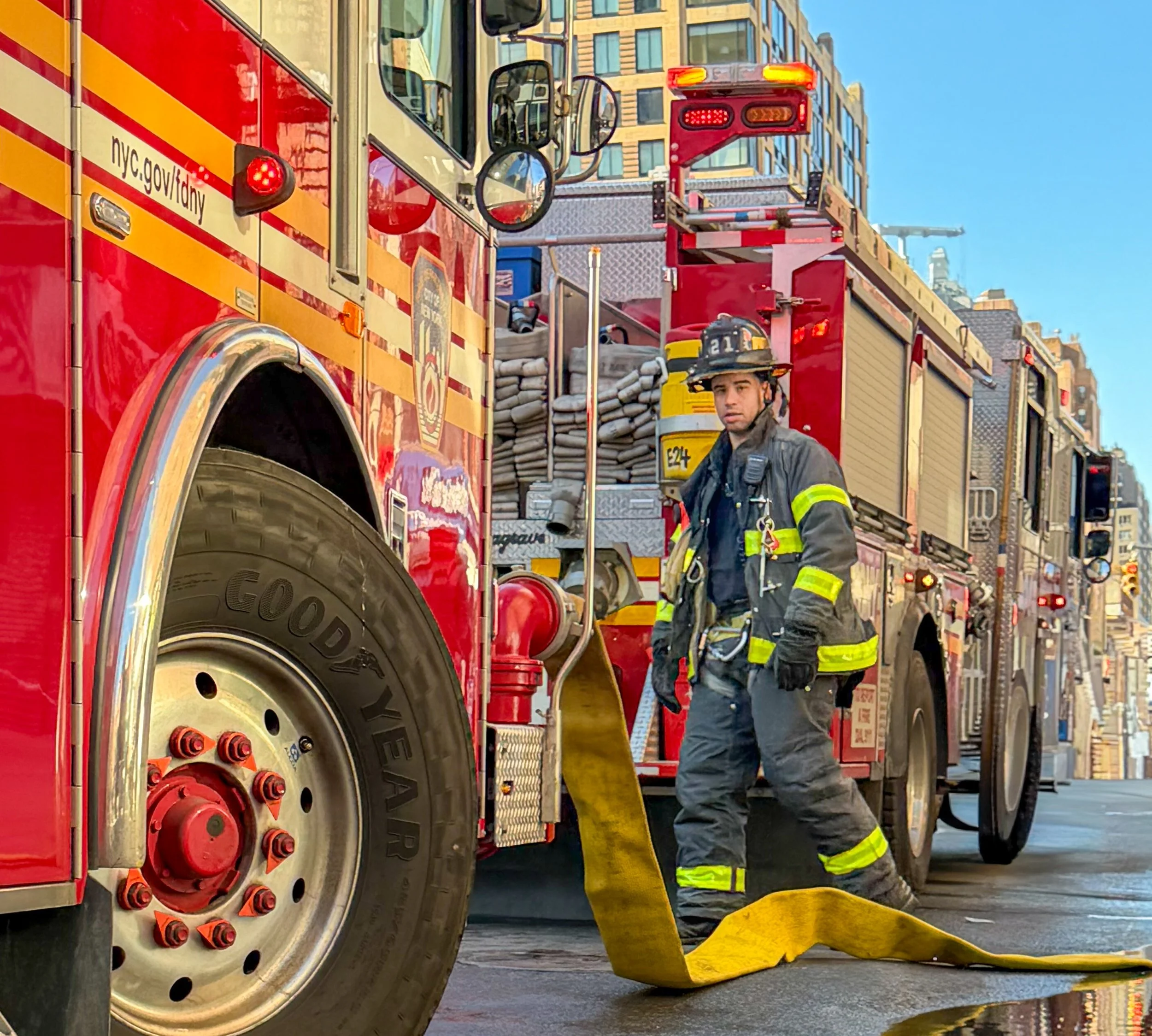 A firefighter in protective gear stands next to a red fire truck while holding a yellow fire hose on a city street.