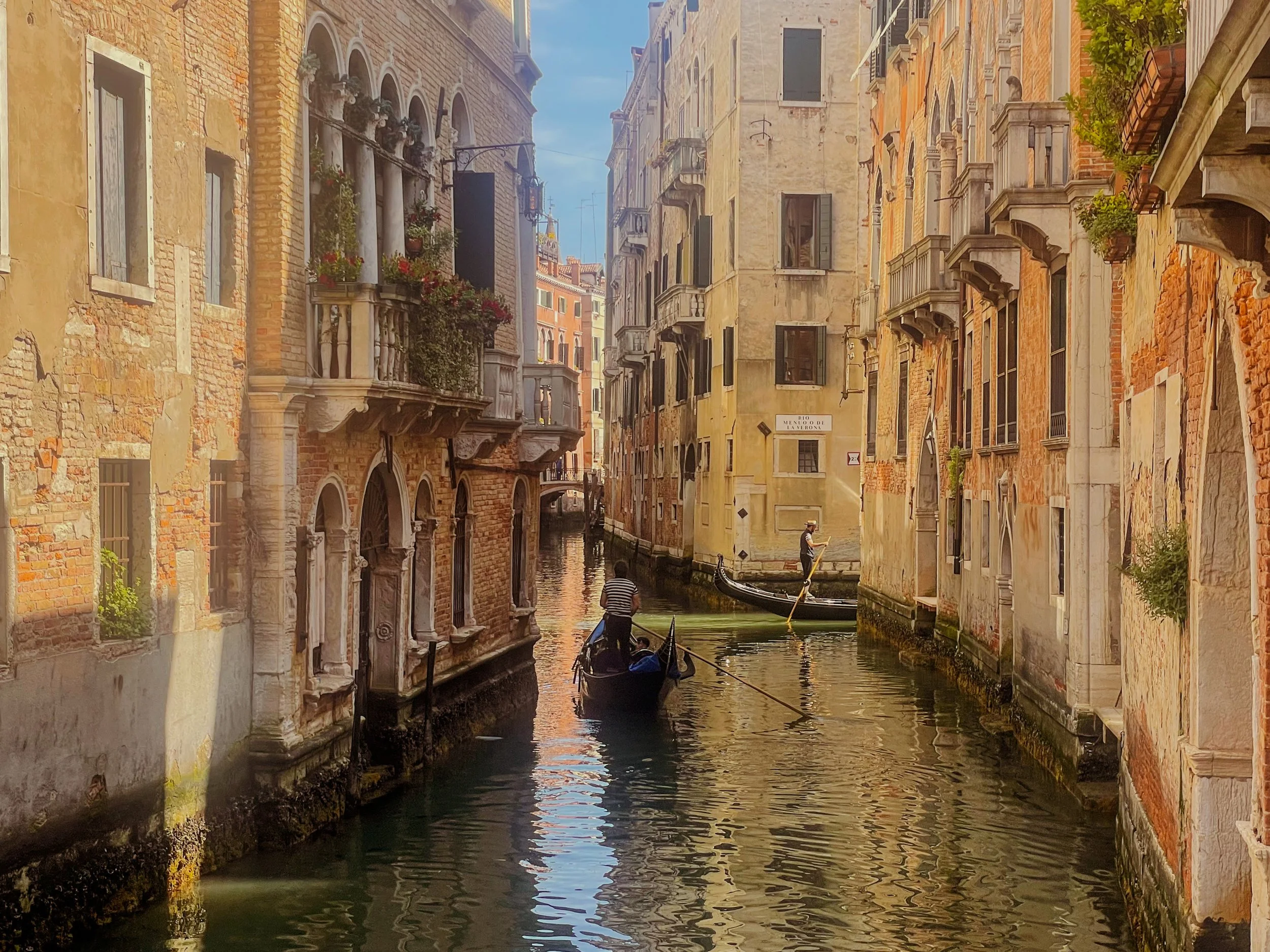 A narrow canal in Venice, Italy, with old brick and stone buildings on either side, and gondolas with gondoliers navigating the waterway.