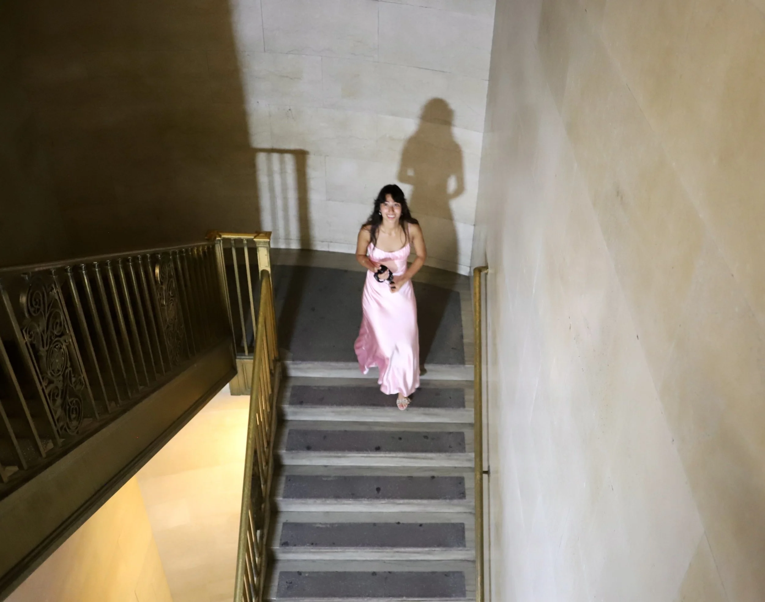 A woman in a pink dress standing at the top of a staircase with a gold railing, smiling and looking up at the camera.