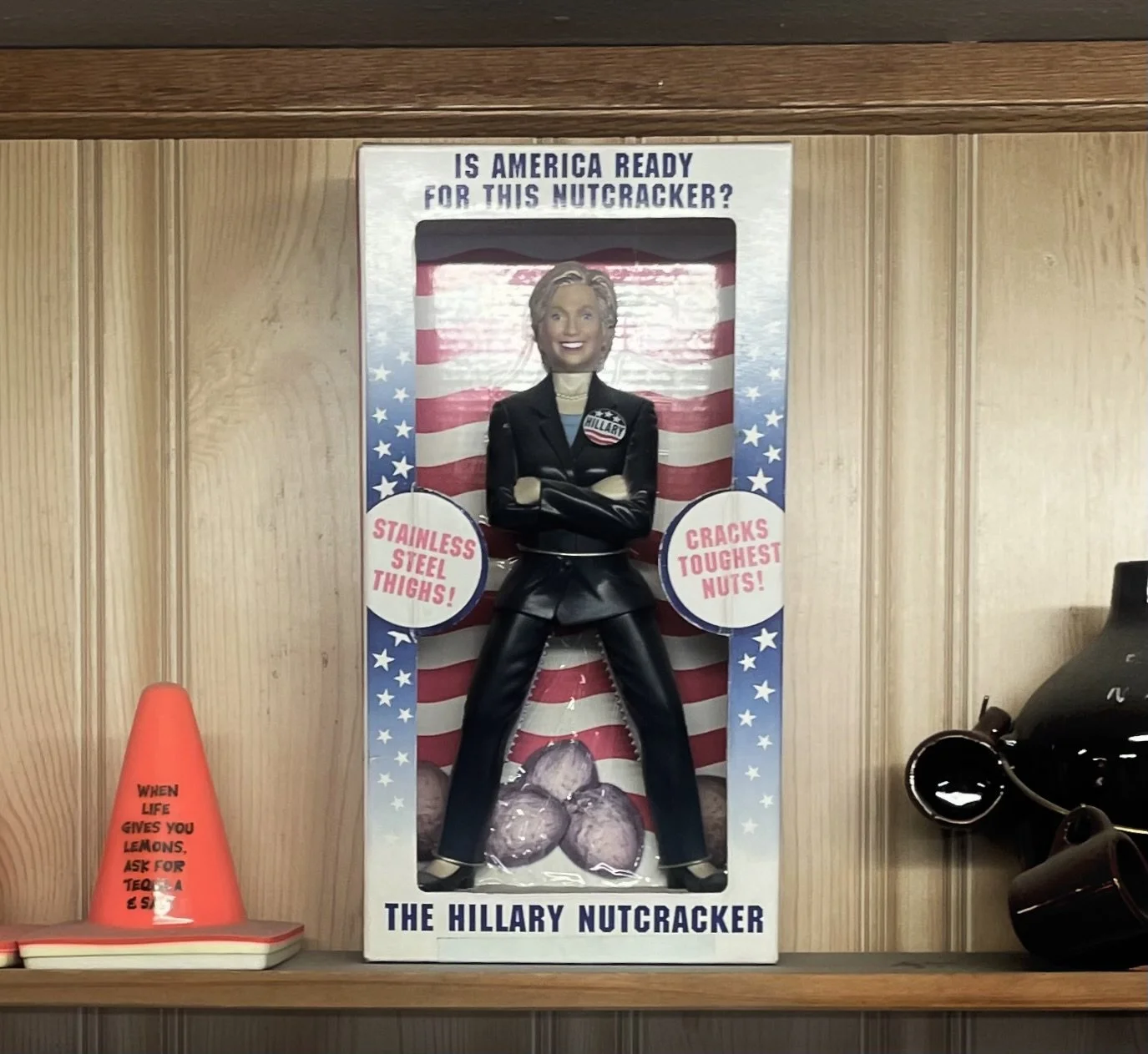 A Hillary Clinton nutcracker with a smiling face, crossed arms, and black leather outfit, standing in front of an American flag background with text promoting its toughness.