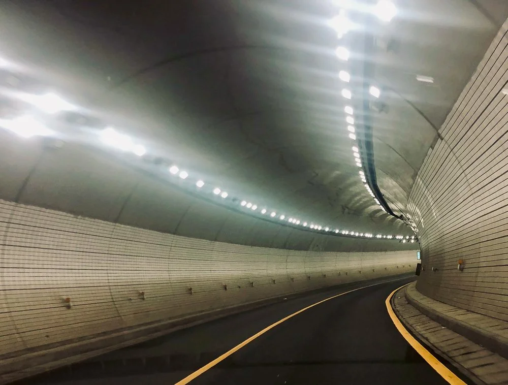 Inside a tunnel with curved walls and ceiling, illuminated by bright overhead lights, and a yellow line marking the edge of the road