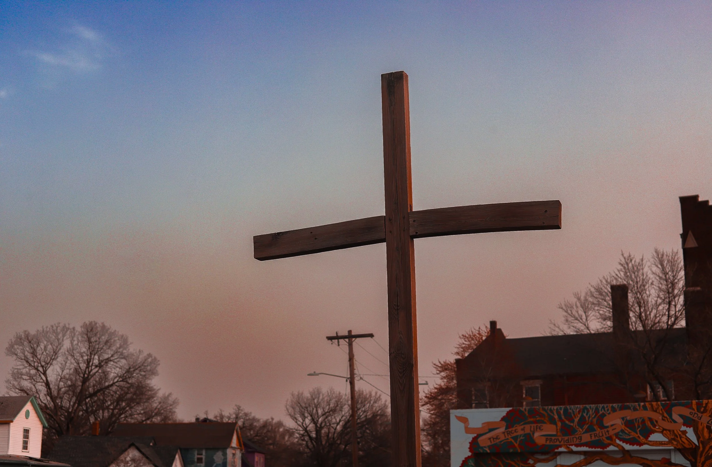 A wooden cross against a sunset sky in a small town with trees and houses.