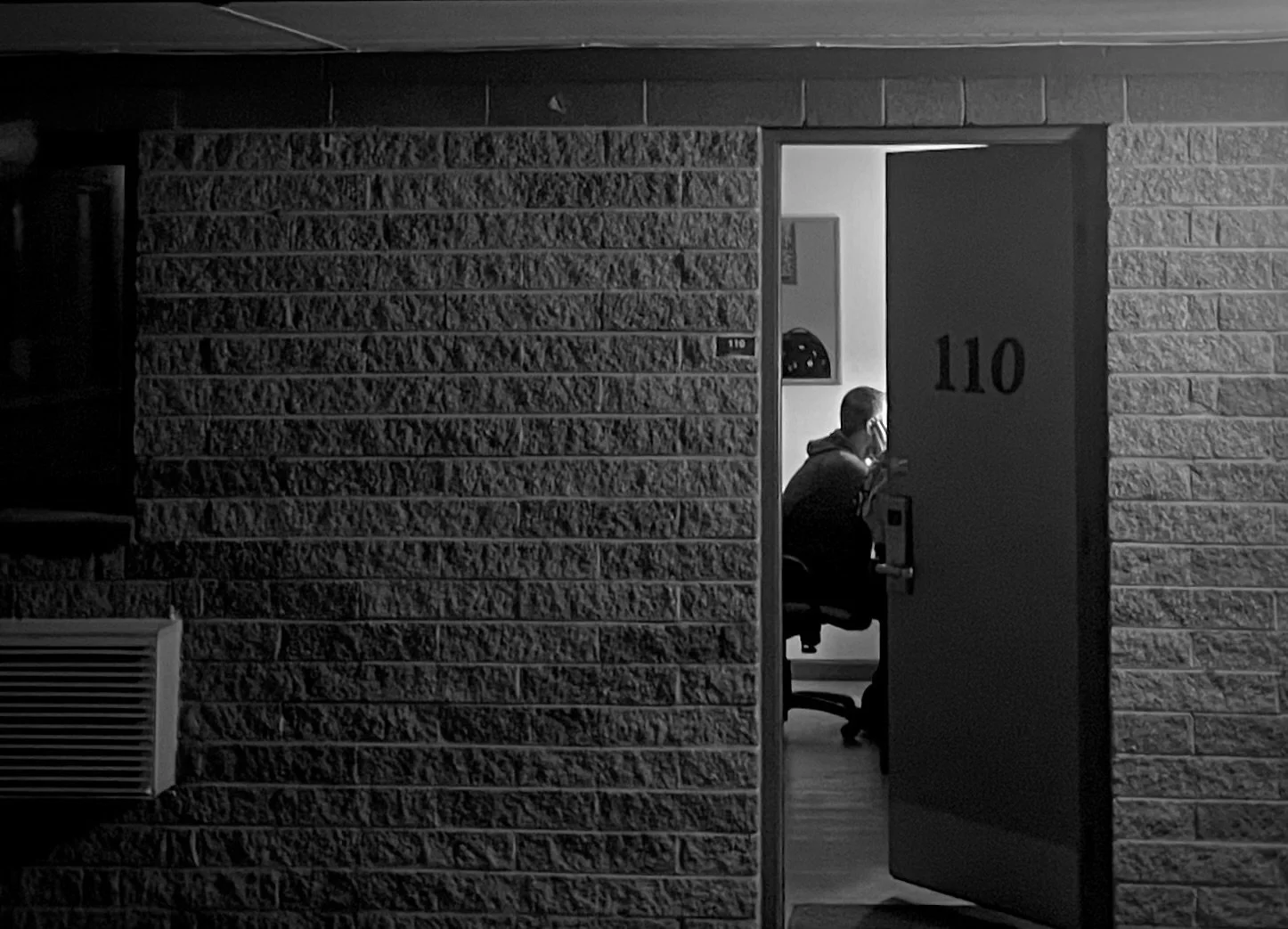 A dimly lit room seen through an open door with the number 110. Inside, a man is sitting at a desk, illuminated by a light, wearing a hoodie, and facing a computer.