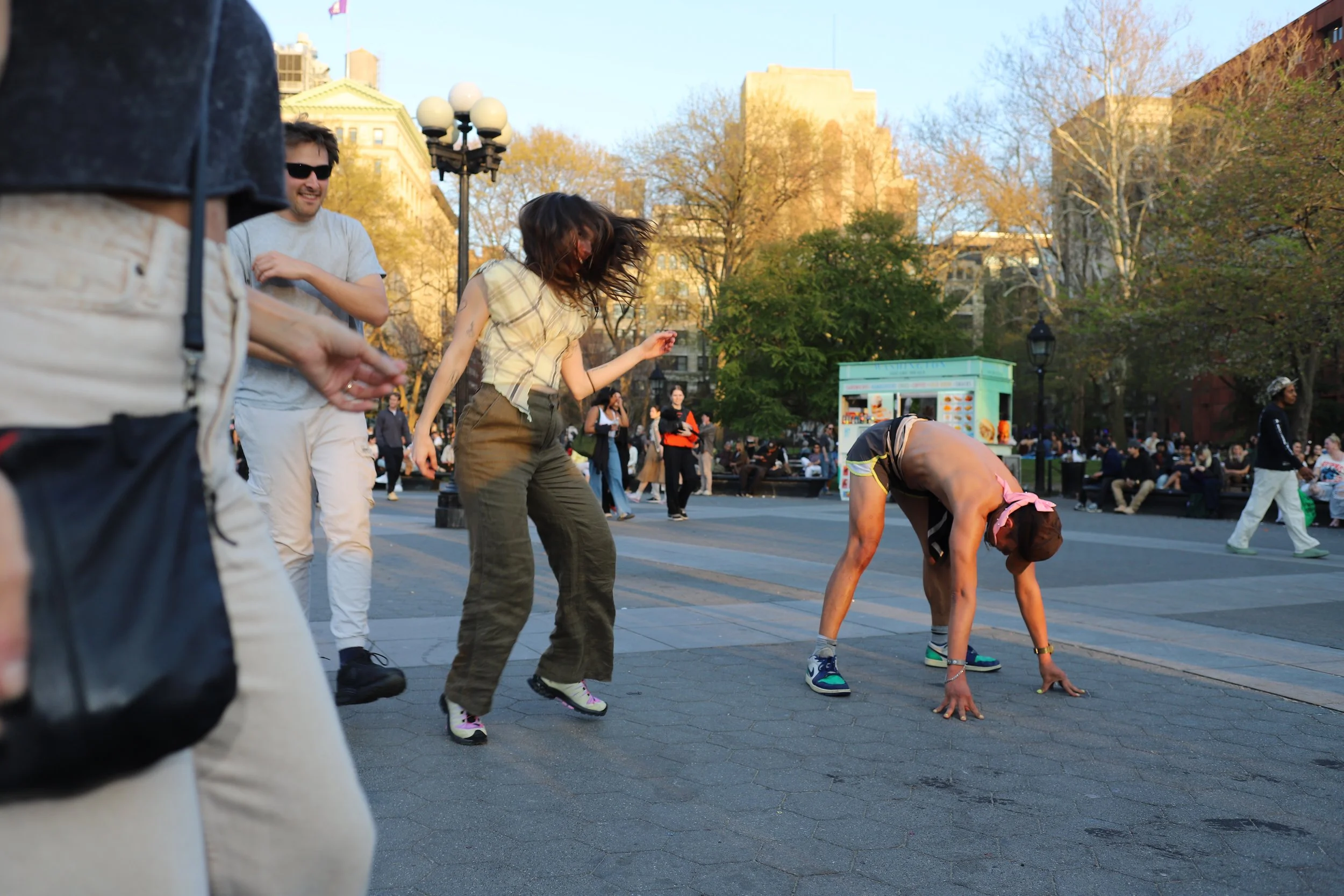People dancing in a park with trees and buildings in the background, some sitting on benches and others walking.