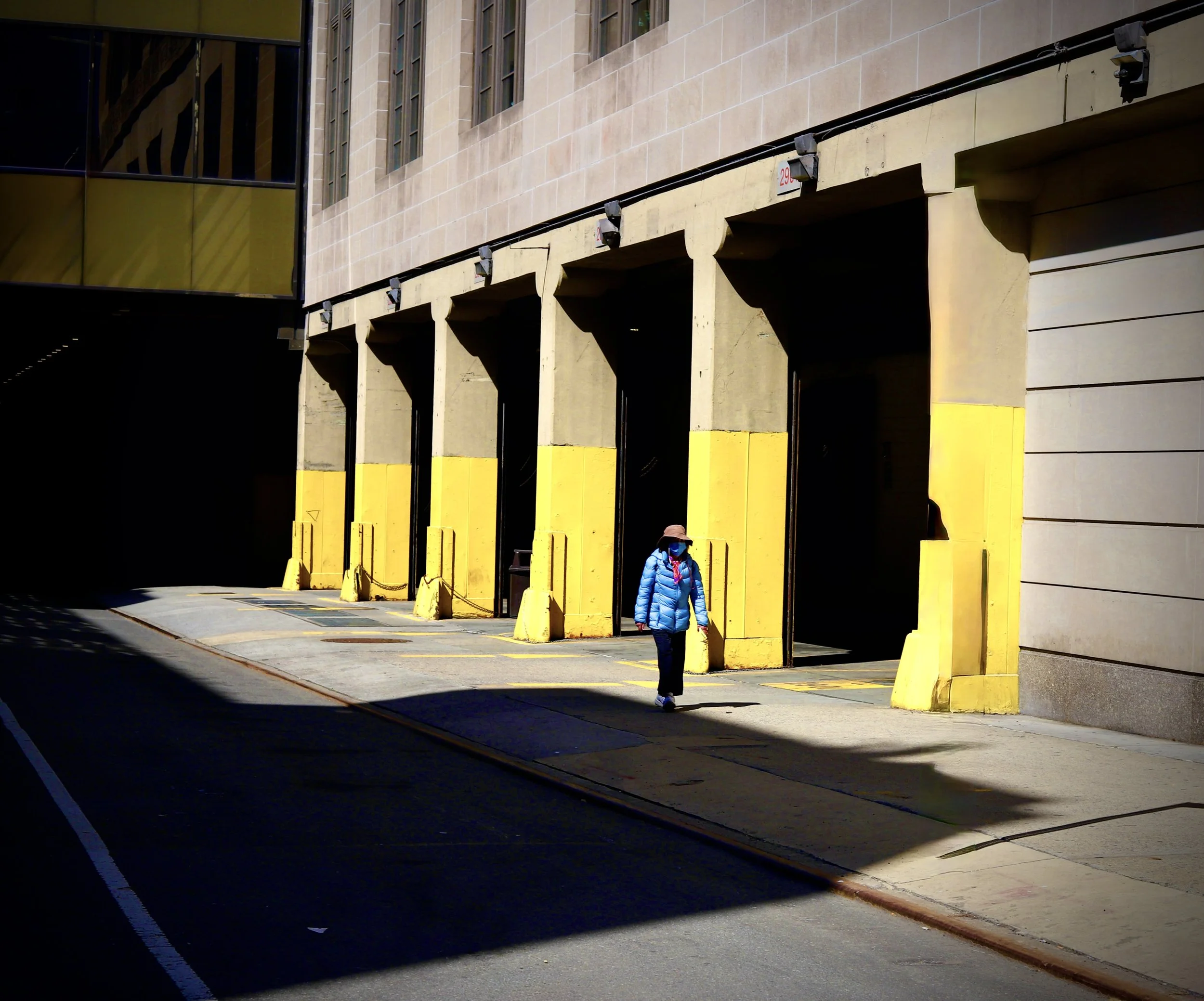 Person wearing a blue jacket, hat, and mask walking past an empty industrial loading dock with yellow protective barriers outside a building.