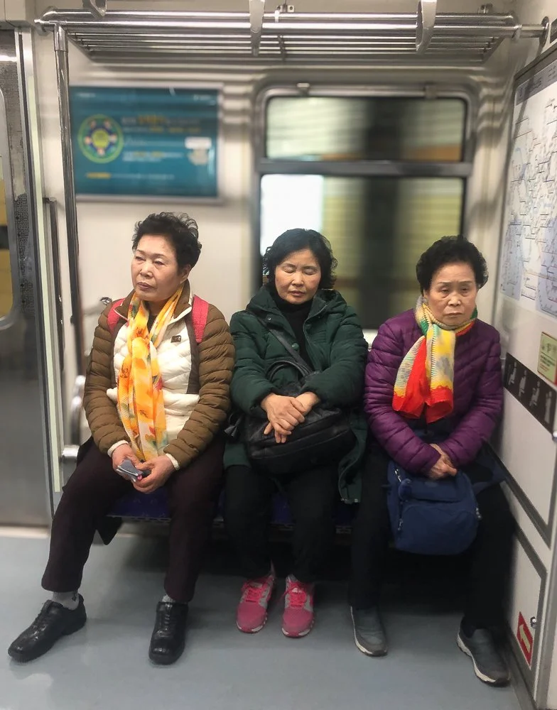 Three women sitting on a train, dressed in jackets and scarves, with closed eyes, appearing tired or asleep.