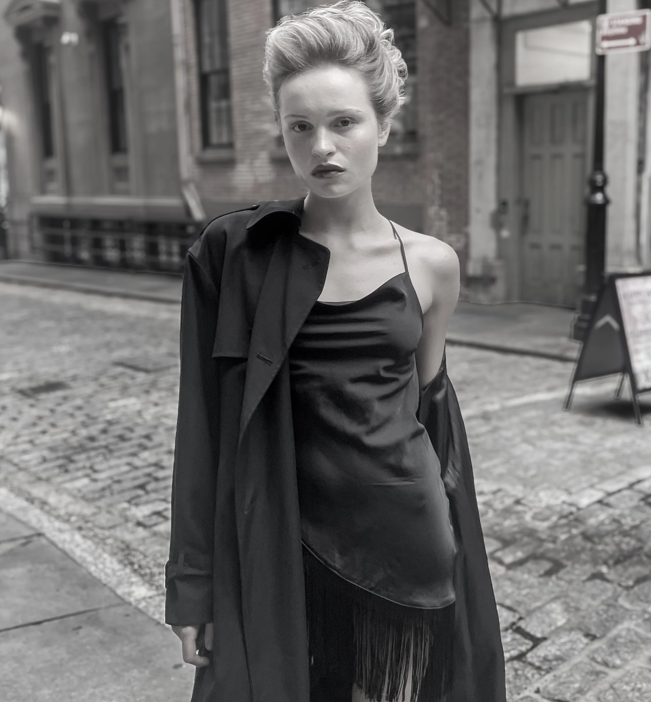Black and white photo of a young woman with short hair in an urban street scene, wearing a satin slip dress and a long coat draped over her shoulders.