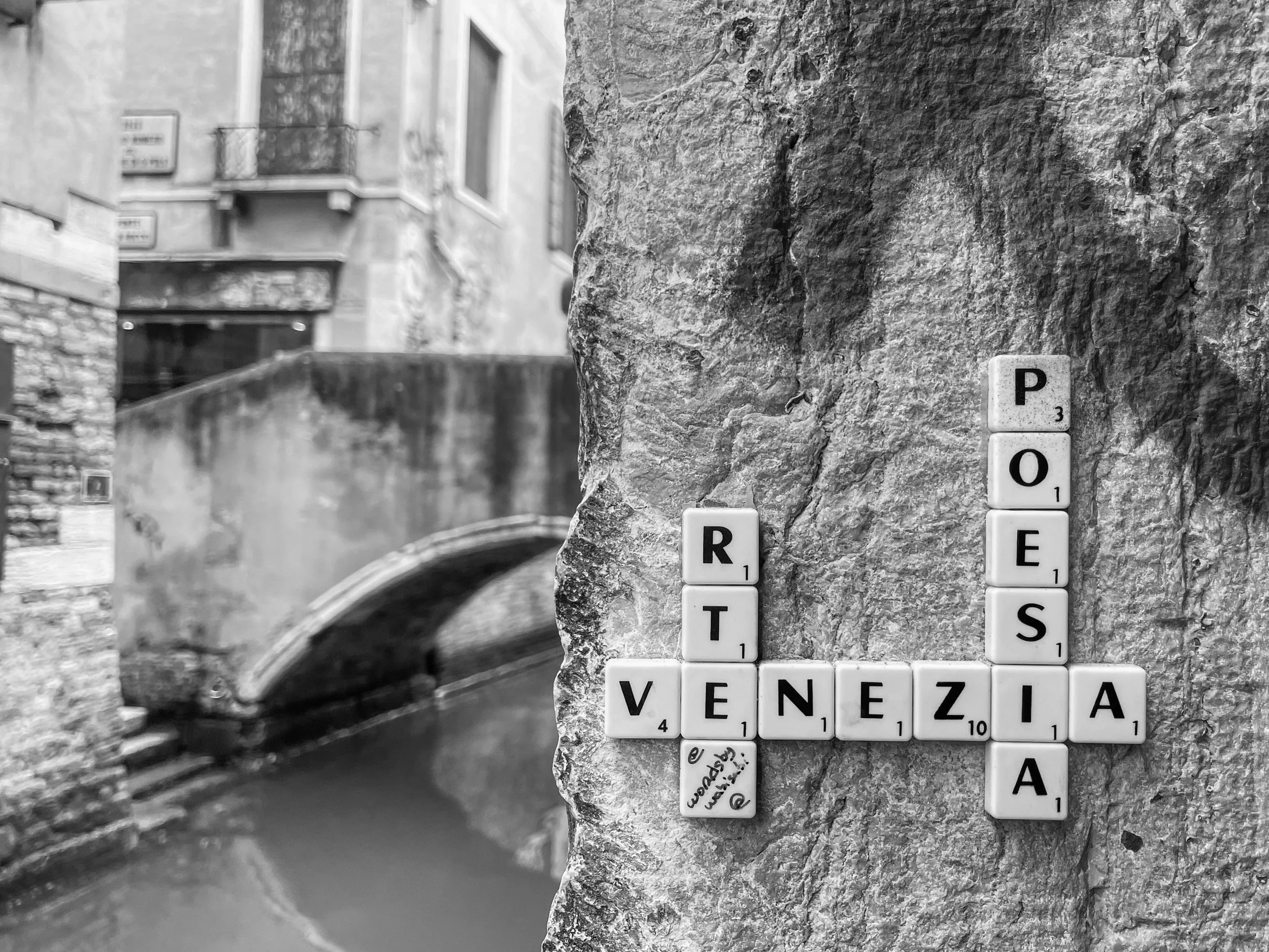 Scrabble tiles spelling out 'VENEZIA' horizontally, with 'RT,' 'PO,' and 'A' vertically. The tiles are attached to a stone wall. In the background, there is a canal with a small stone bridge over it, surrounded by old buildings, some with balconies.