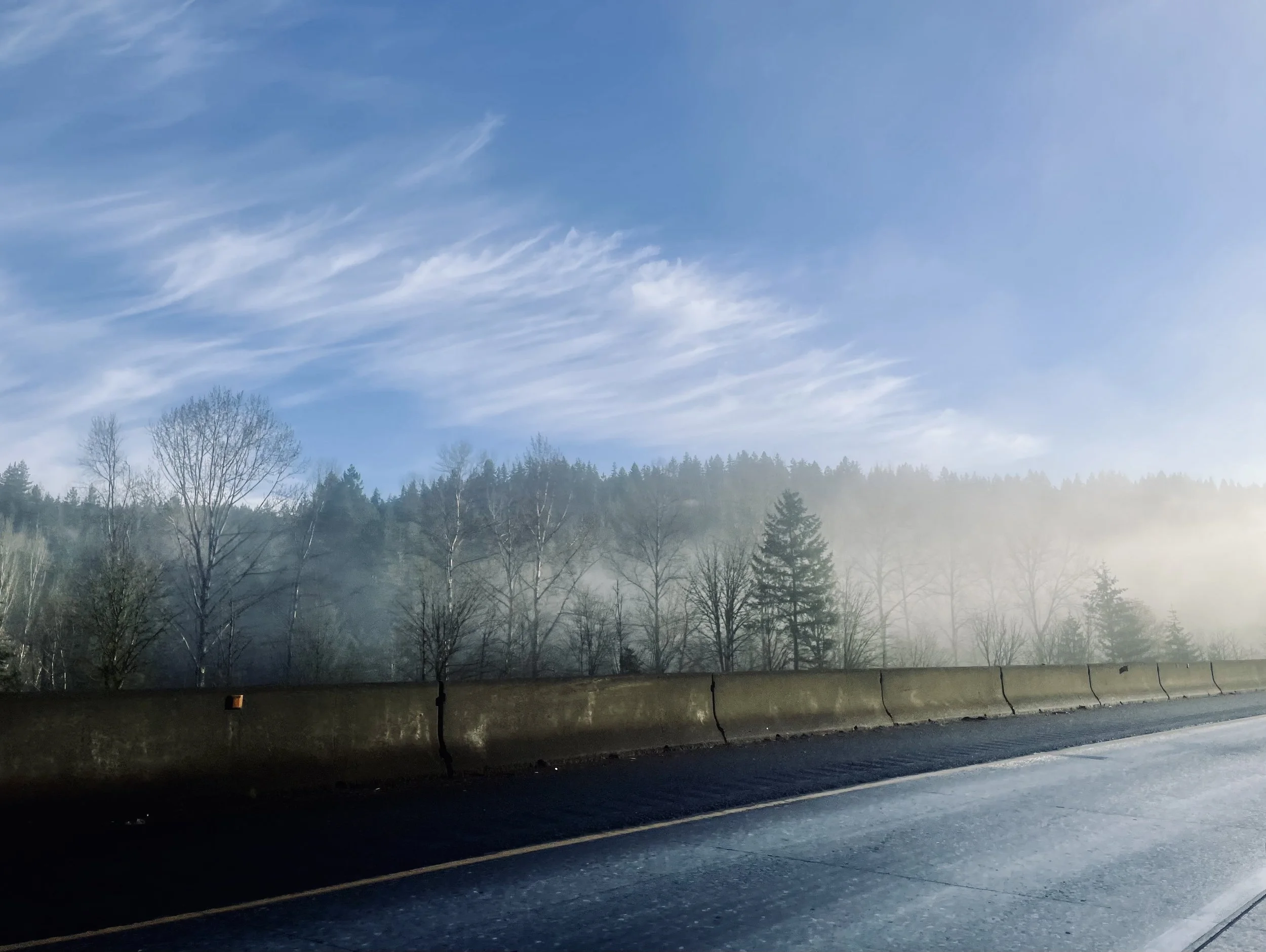 A highway with a foggy forest in the background and a blue sky with wispy clouds.