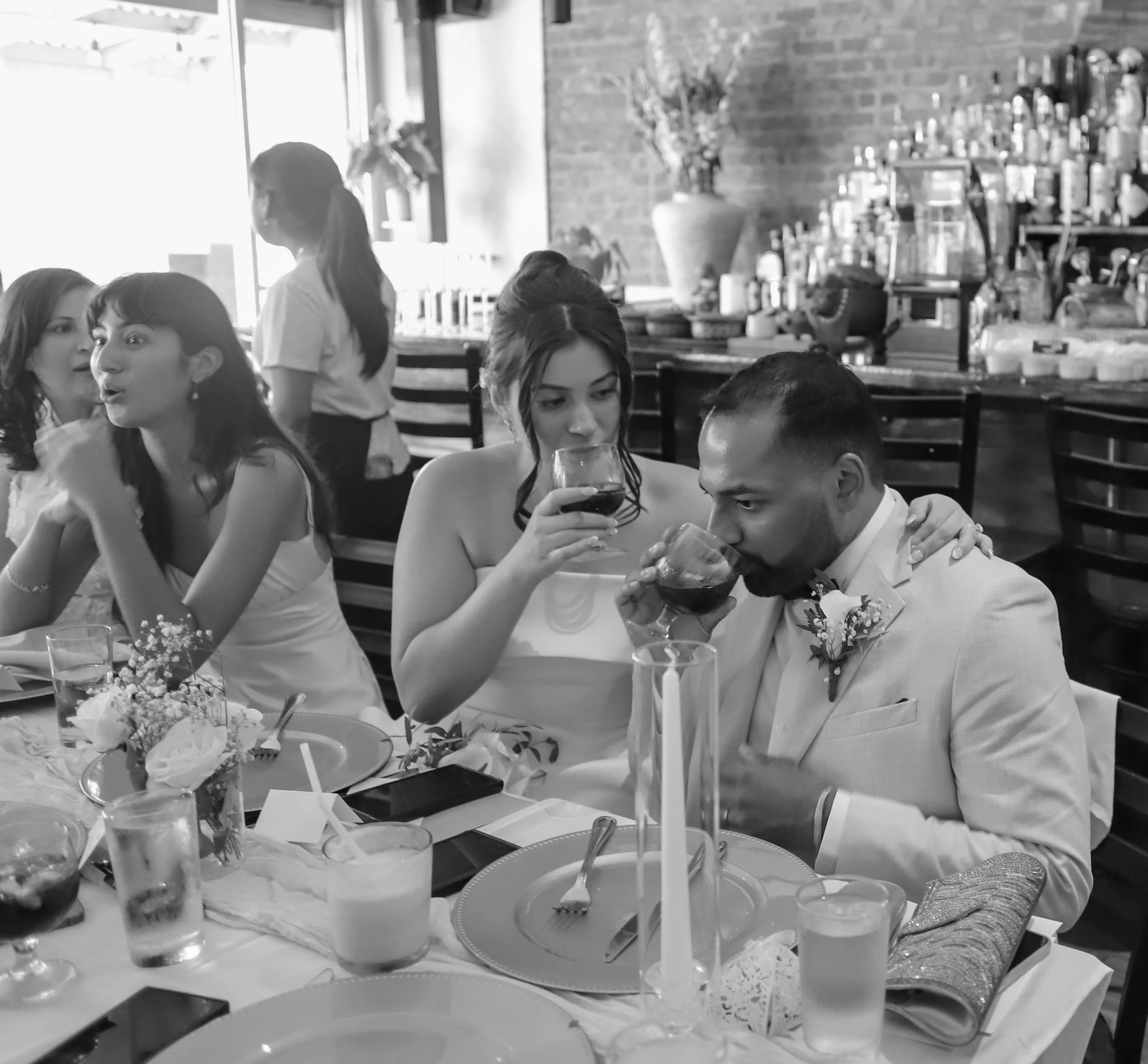 People celebrating at a wedding reception, with a woman drinking wine and a man in a suit sipping from a glass, surrounded by guests and table settings in a restaurant or banquet hall.