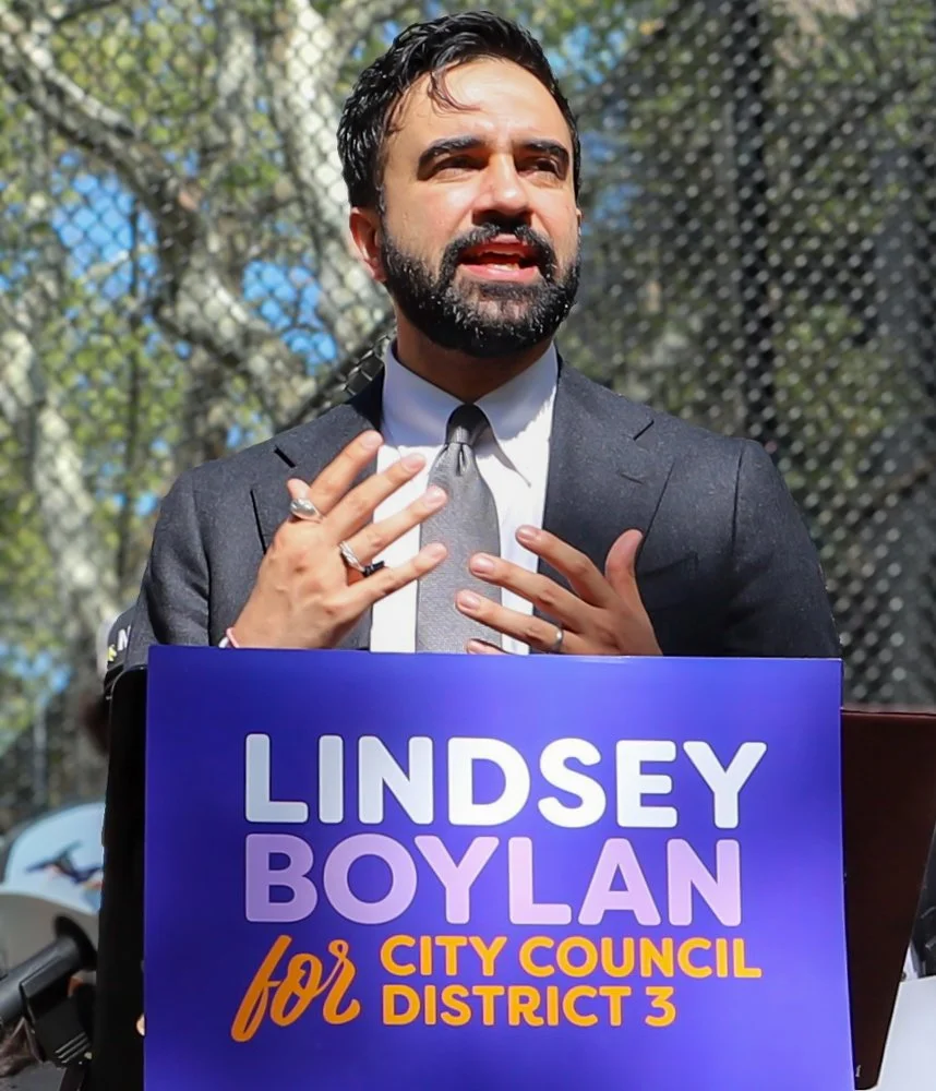 Man in a suit speaking at a rally, holding his chest with both hands, behind a sign that reads 'Lindsey Boylan for City Council District 3' with a blue background.