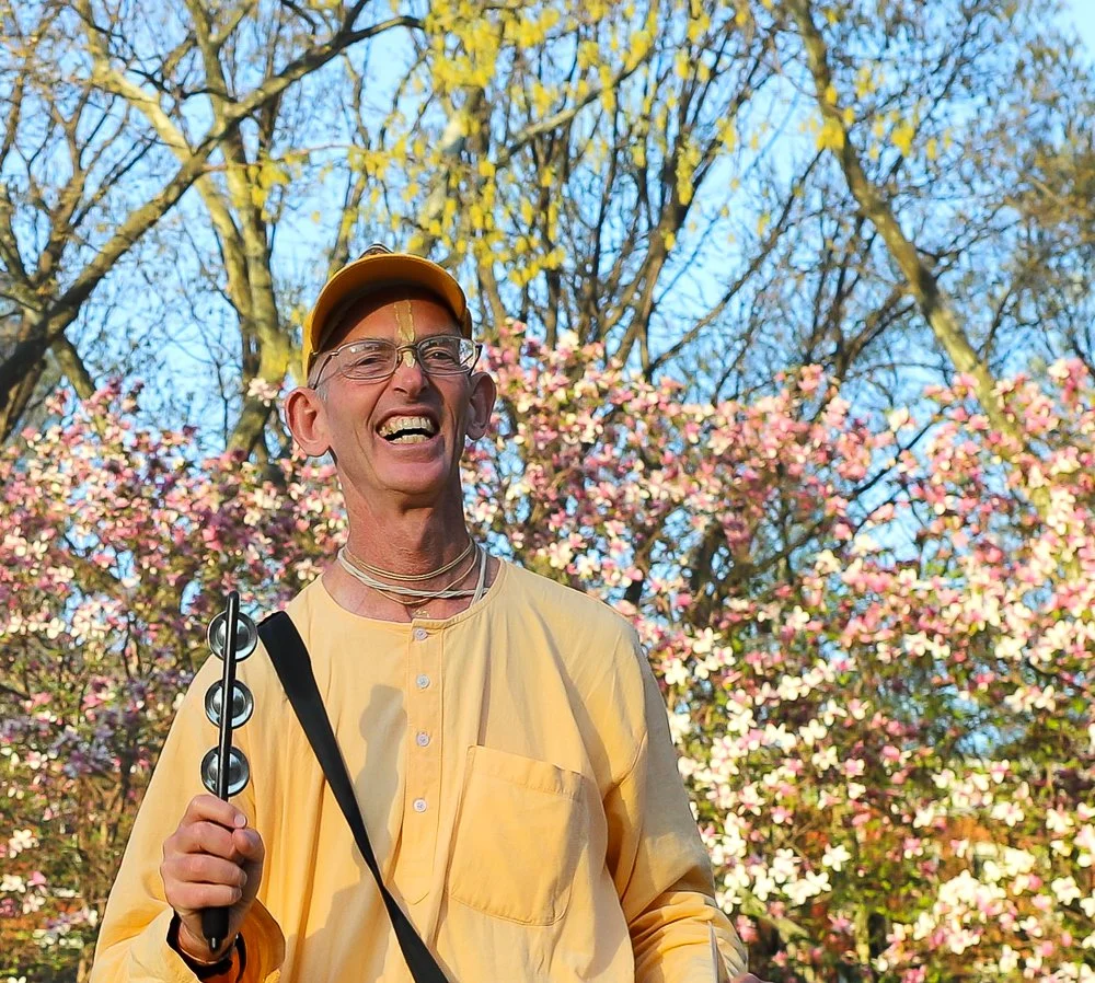 A smiling man wearing glasses, a yellow cap, and a yellow shirt, holding a musical instrument, standing outdoors in front of blooming pink flowers and leafless trees under a blue sky.