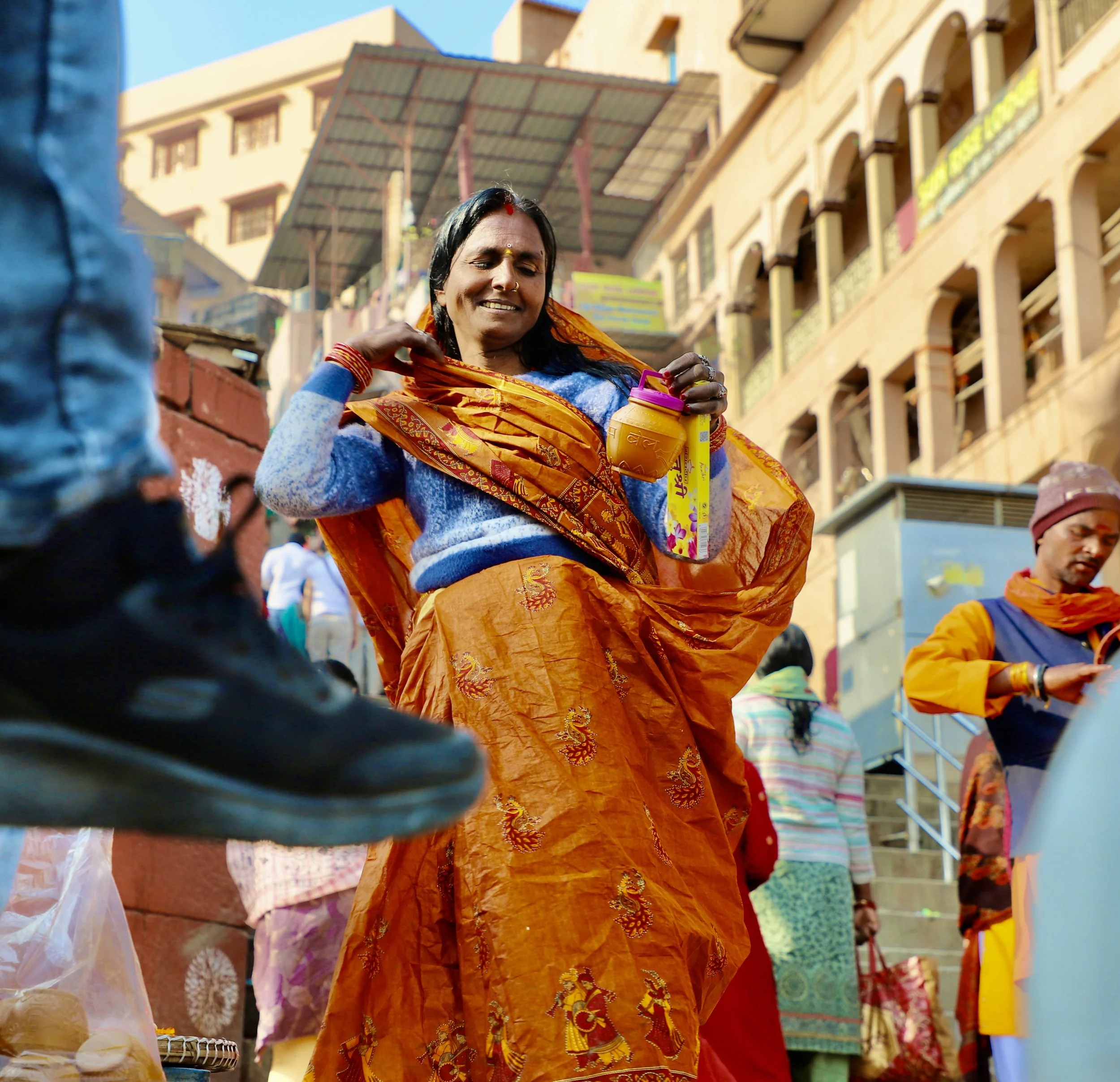 A smiling woman in traditional orange and blue attire celebrating in a street market in India, holding a colorful container, with crowded buildings and other people in the background.