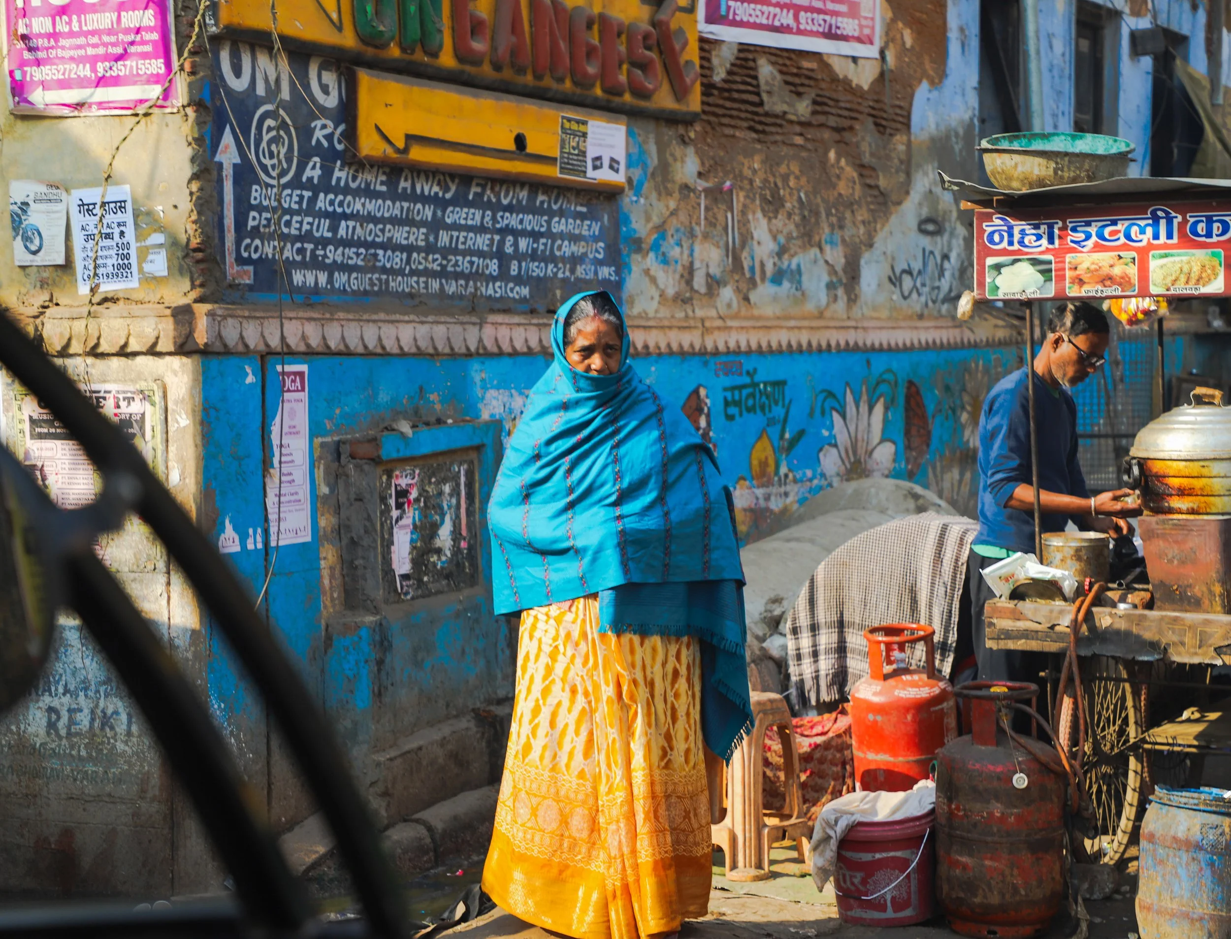 A woman wearing a blue shawl and yellow skirt standing on a street next to a man cooking at a street food stall with gas cylinders and cooking utensils, in front of a painted wall with signs and posters.