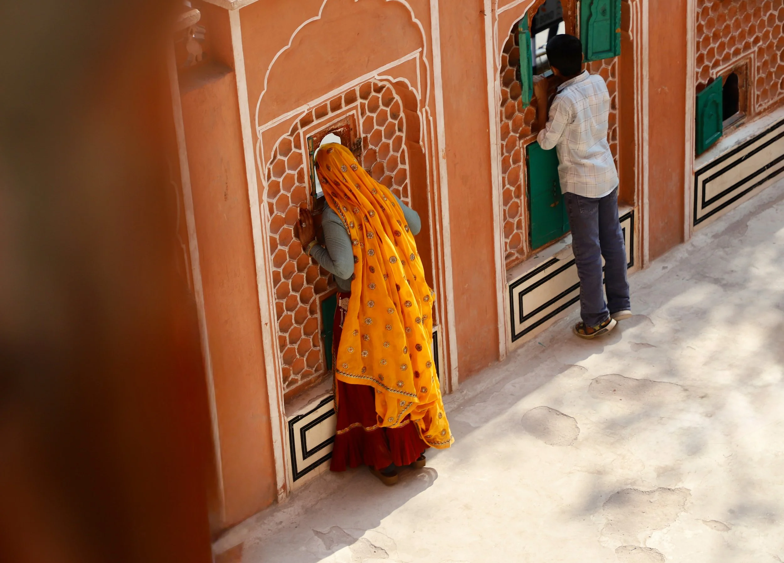 A woman in a yellow traditional dress and head covering is praying at a small window of a pinkish-orange wall, with a man in a checked shirt and jeans praying at a similar window nearby.