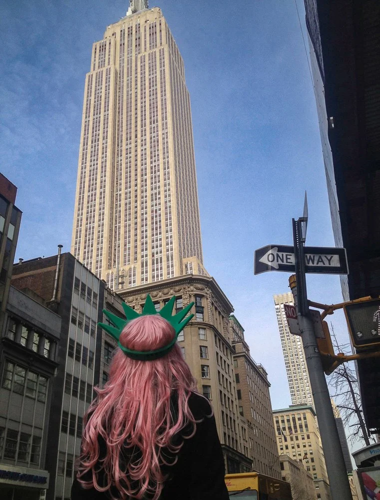 A person with long pink hair wearing a green Statue of Liberty headband standing on a city street in front of tall buildings and a blue sky.