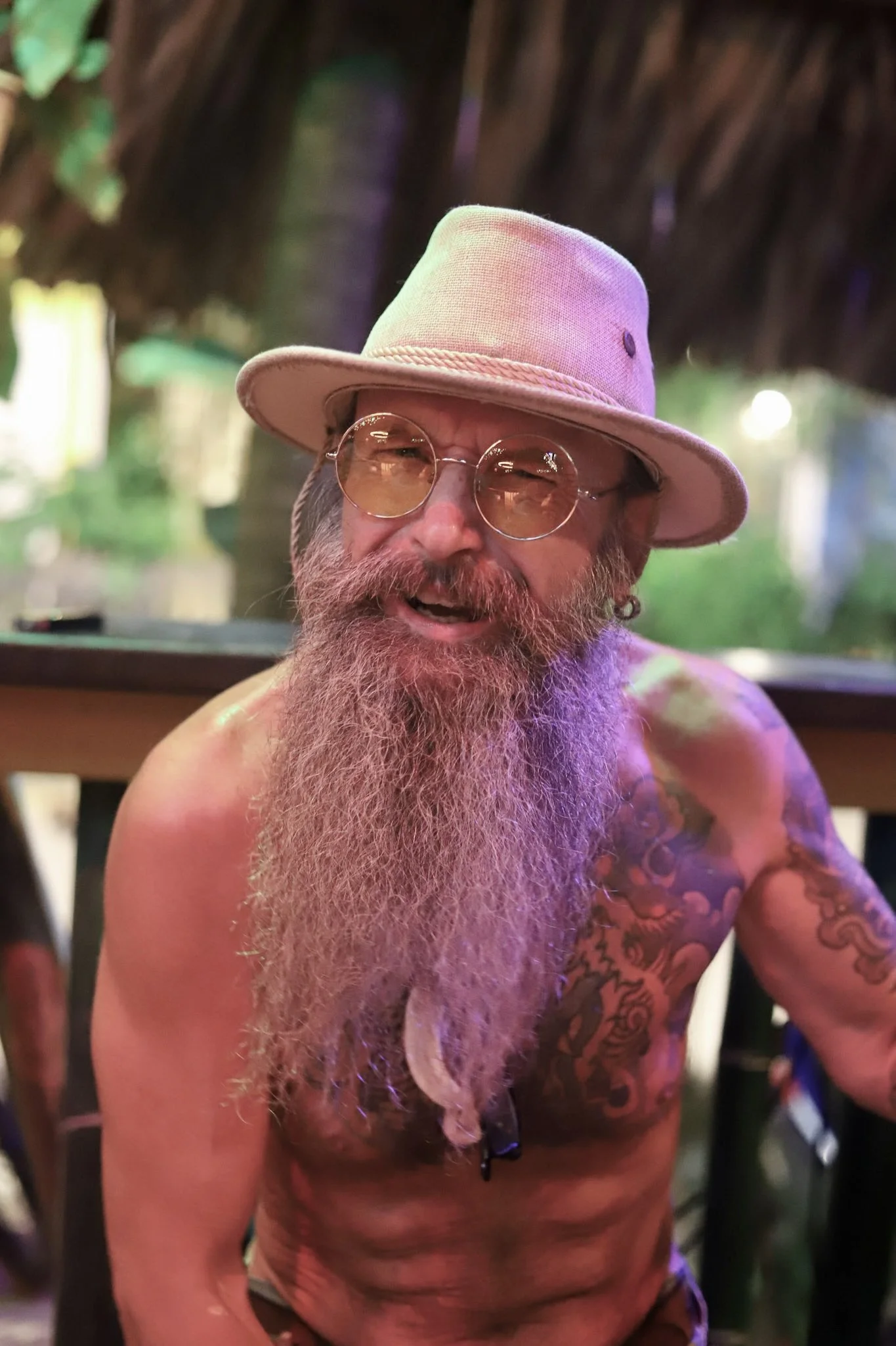 A shirtless man with a long grey beard, glasses, a light-colored hat, and tattoos on his shoulders, sitting outdoors under a thatched roof, smiling at the camera. Roatan