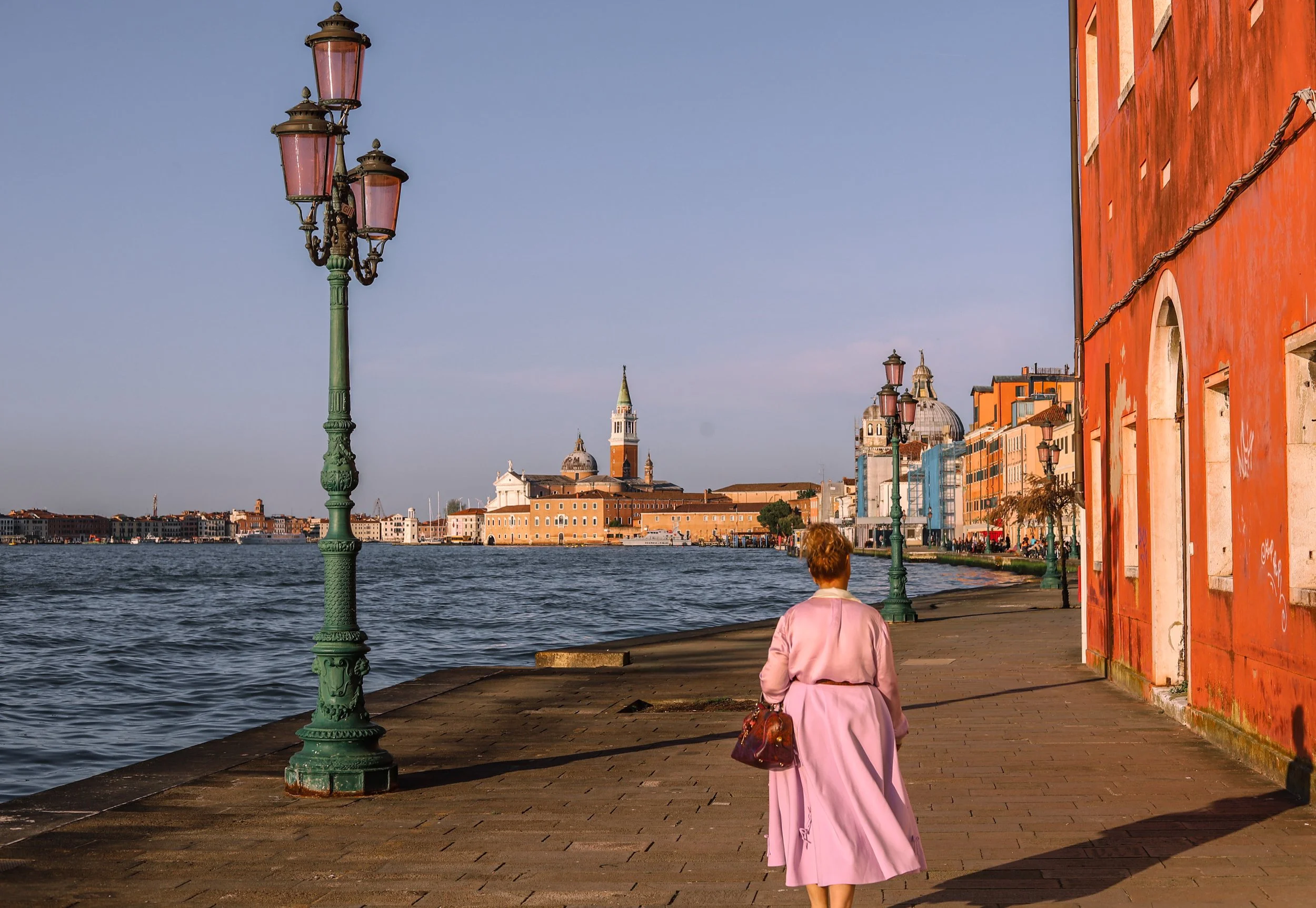 A woman in a pink coat walking along a waterfront promenade with lamp posts in Venice, Italy, with historic buildings and the water in the background.