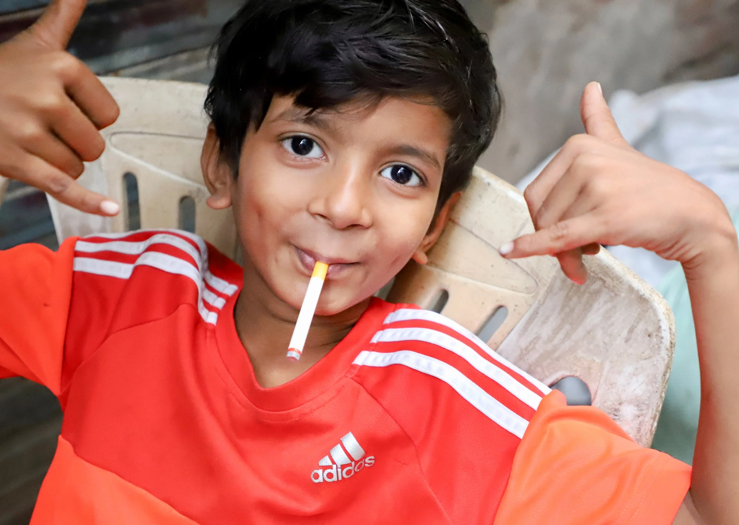 A young boy with dark hair and brown skin is smiling and pointing at a cigarette in his mouth. He is wearing an orange Adidas sports shirt with white stripes on the sleeves, and he is sitting on a beige plastic chair.
