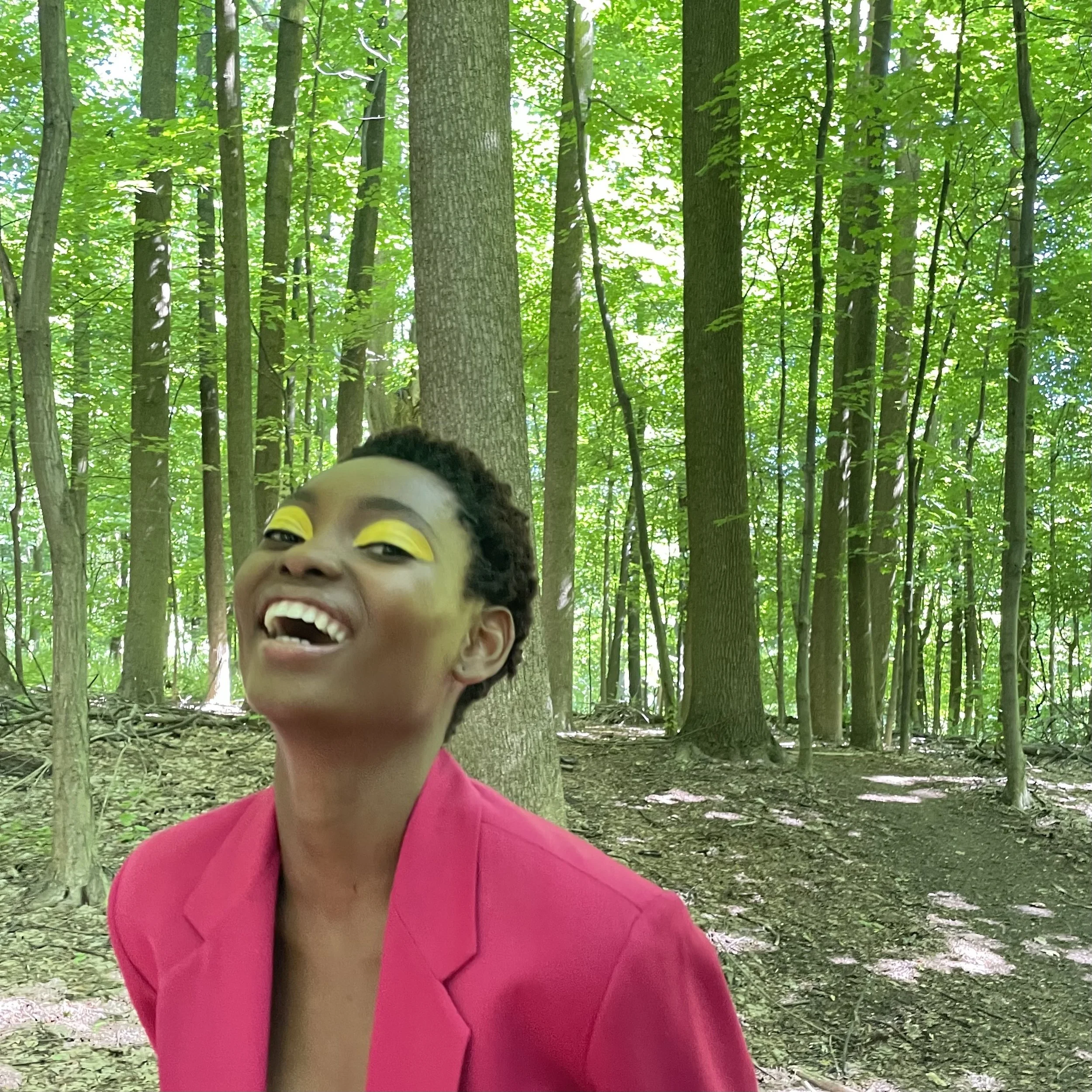 A woman with short curly hair and wearing a bright pink blazer, standing in a lush green forest with tall trees and sunlight filtering through the leaves. She has vibrant yellow eyeshadow and is smiling joyfully.