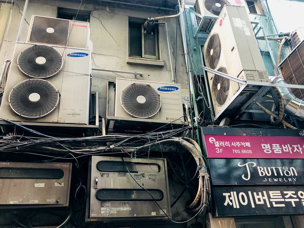 Multiple air conditioning units mounted on an urban building exterior with a network of wires and cables beneath them. Signs in Korean and English are visible on a shop below.