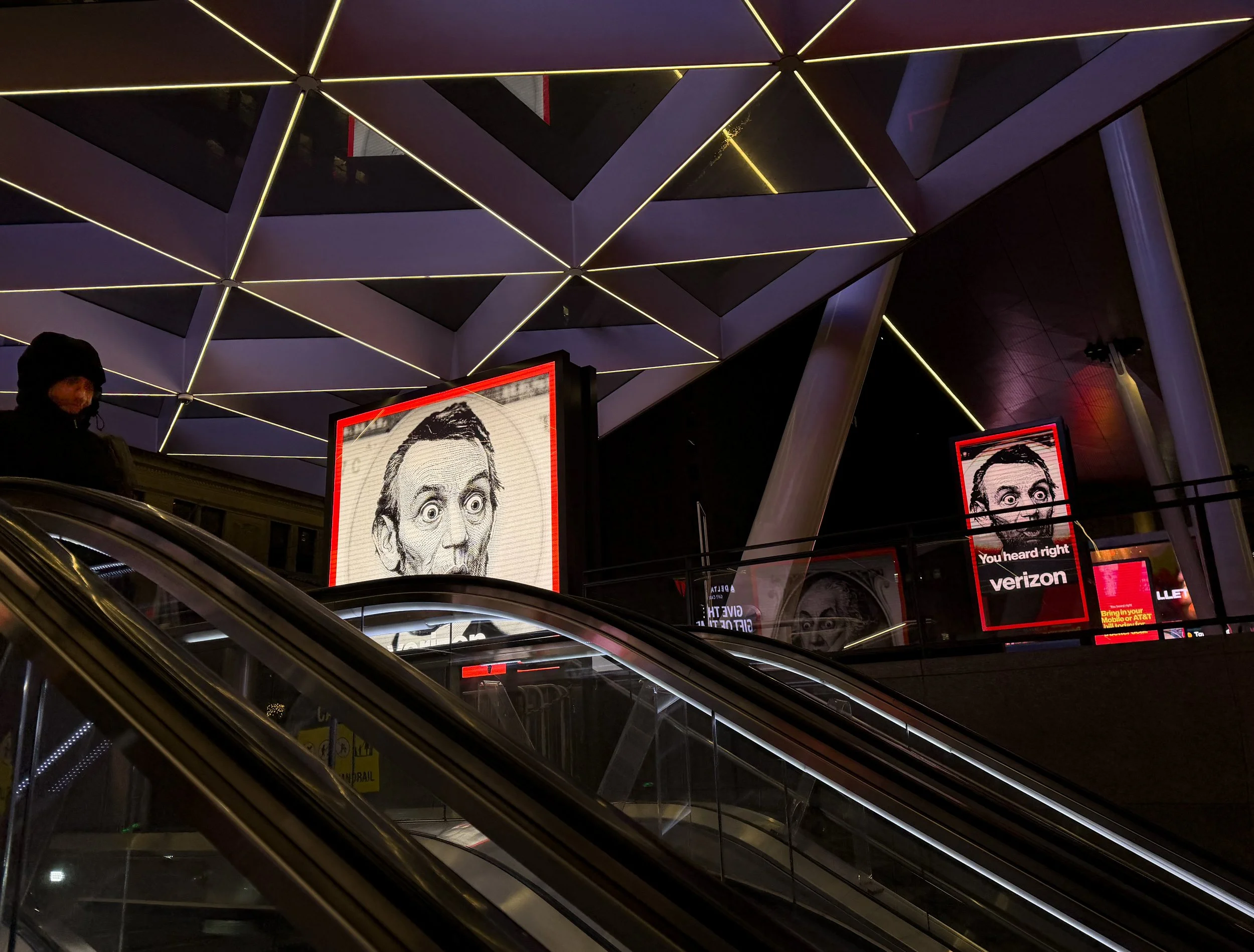 Nighttime scene at a modern indoor space with escalators, illuminated geometric ceiling designs, and multiple large digital screens displaying black and white images of a man's face with wide eyes and exaggerated expression.