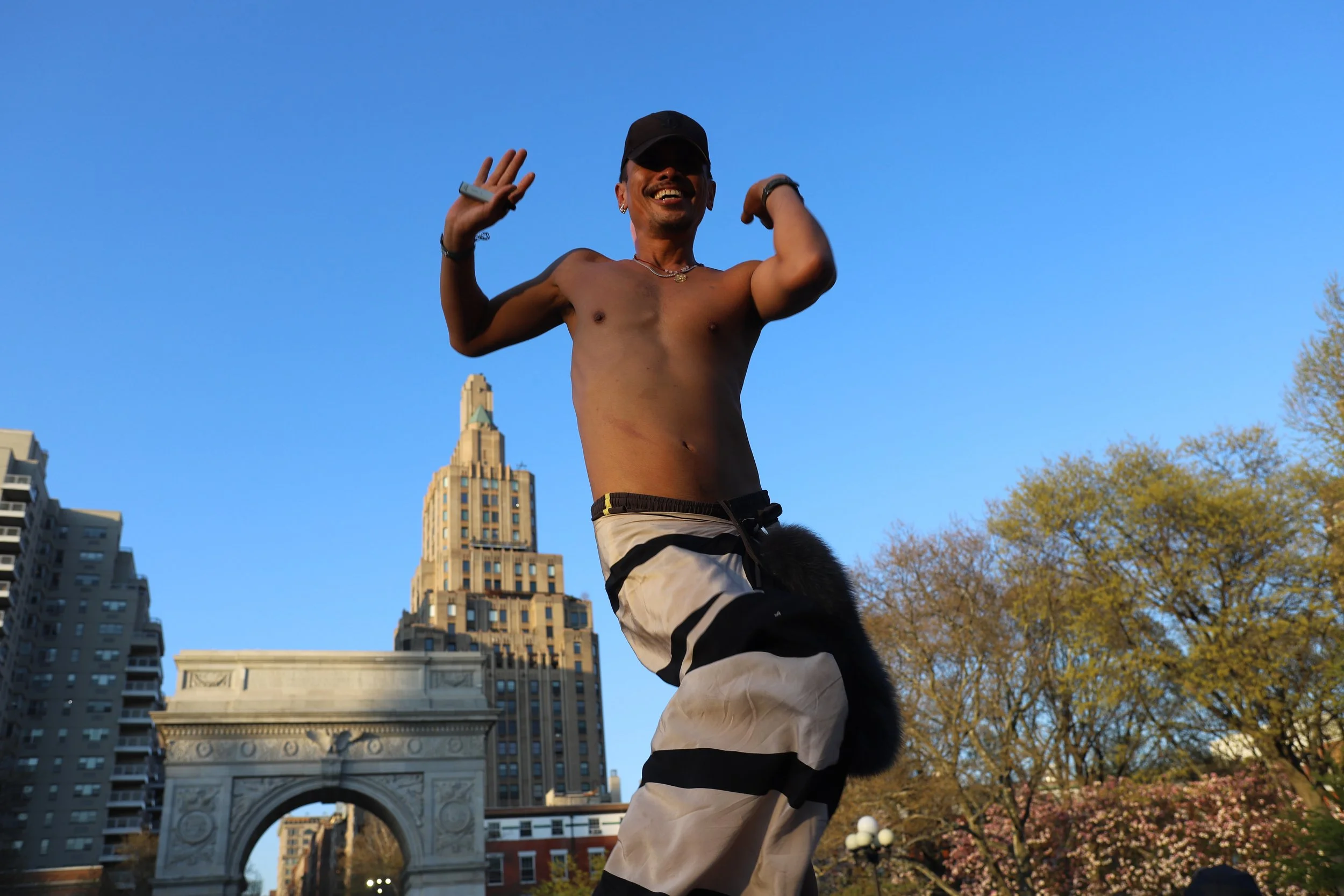 A shirtless man wearing a black cap and striped shorts, smiling and posing with arms raised, stands outdoors with city buildings and trees in the background.