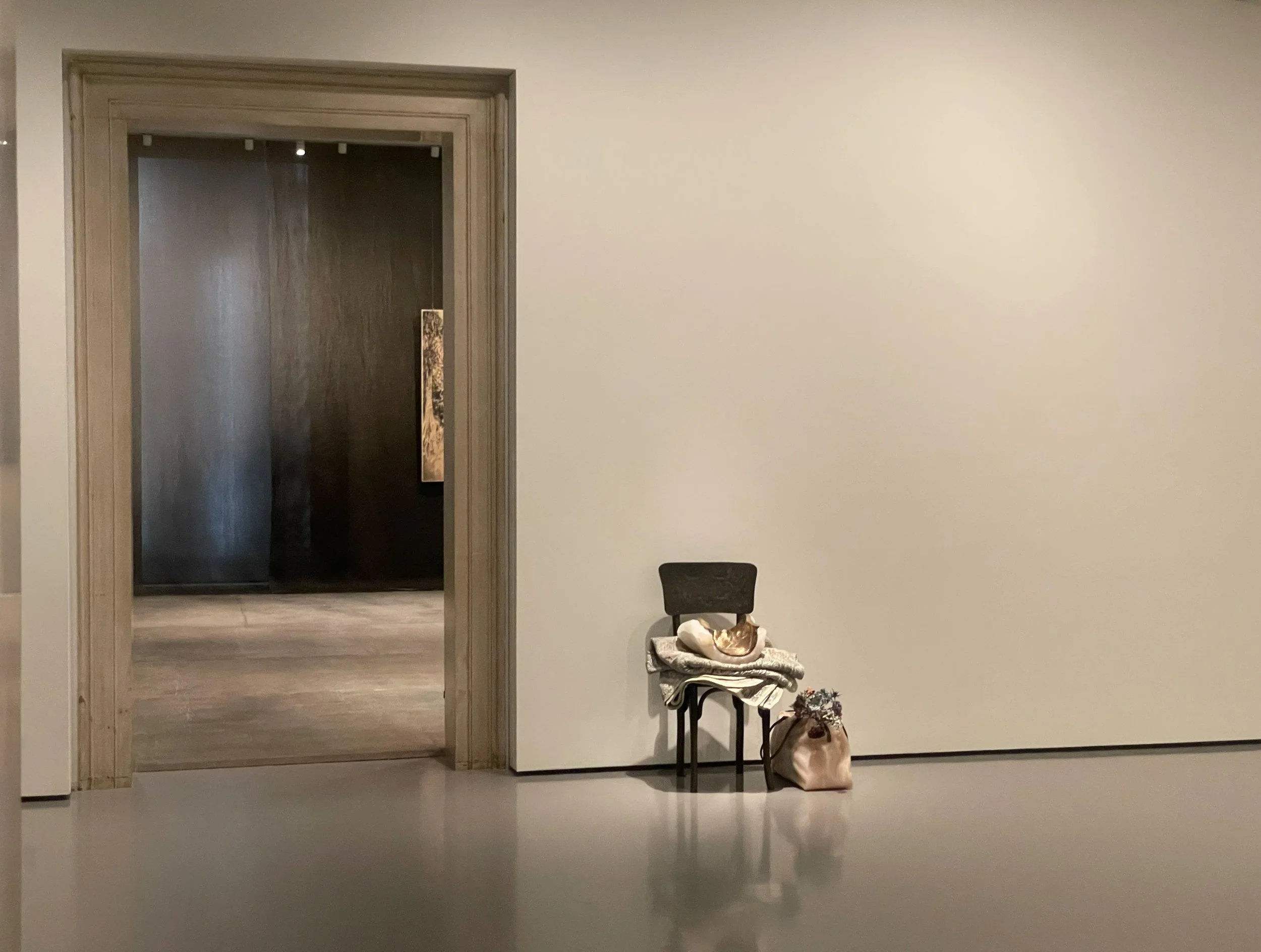 A black chair with a cloth and a bag hanging on it, a cat curled up sleeping on a blanket on the chair, and a bag with flowers on the ground beside the chair, against a plain white wall with an open doorway leading to a darker room.