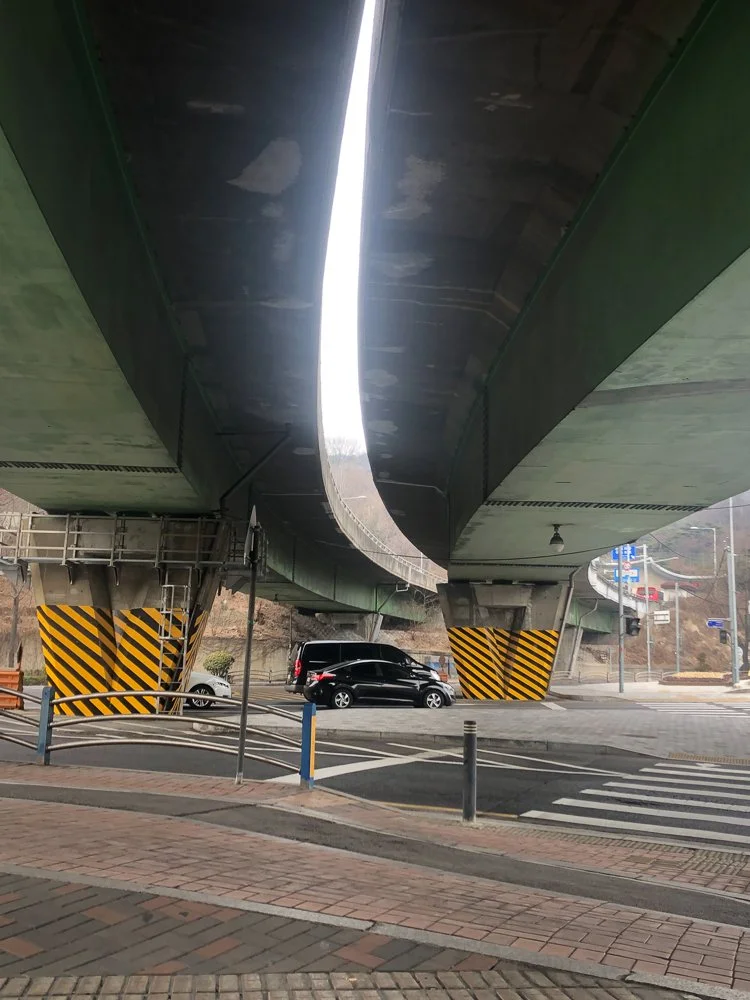 Underneath a large highway overpass with multiple cars parked below, featuring black and yellow striped safety markings on the supporting columns, and a crosswalk on the street.