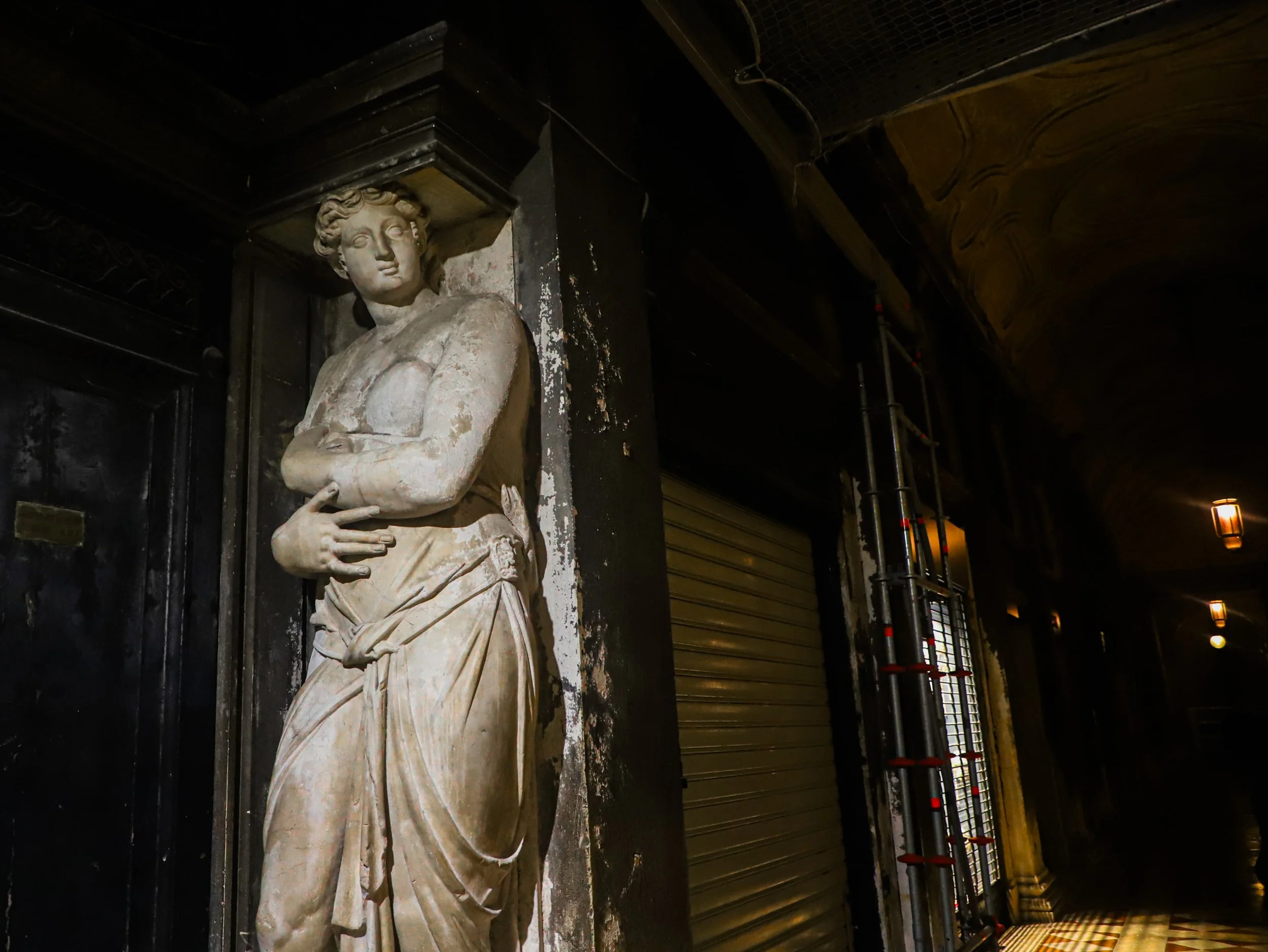 An illuminated classical statue of a woman with arms crossed, standing next to a dark door in a dim hallway with arched ceiling and wall-mounted lights.