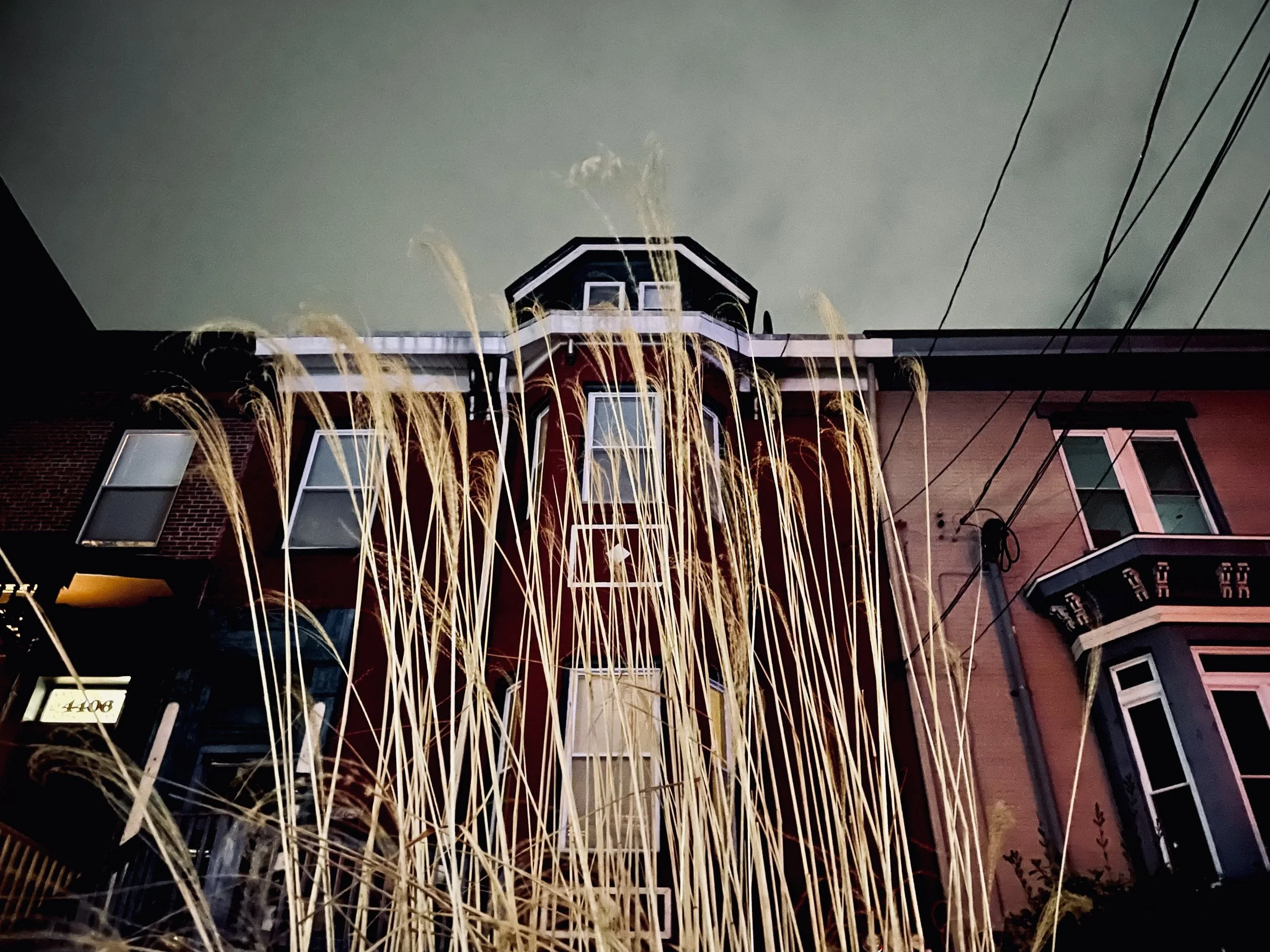 Nighttime photo of old Victorian-style row house with tall grass in foreground, power lines overhead, and dark cloudy sky.