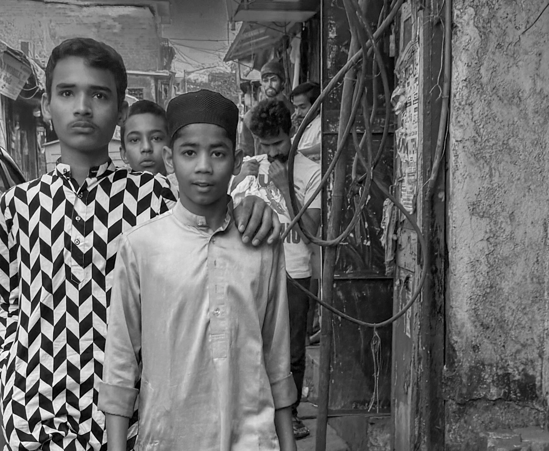 Four young boys standing in a street with older boys in the background among small shops and market stalls.