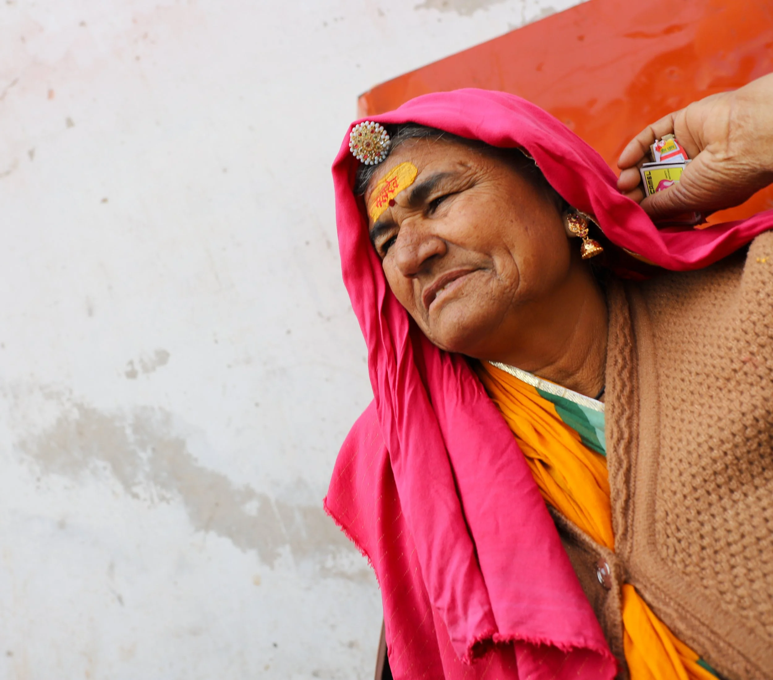 An elderly woman dressed in traditional colorful attire with gold jewelry and a pink headscarf, receiving a small item from a person's hand.