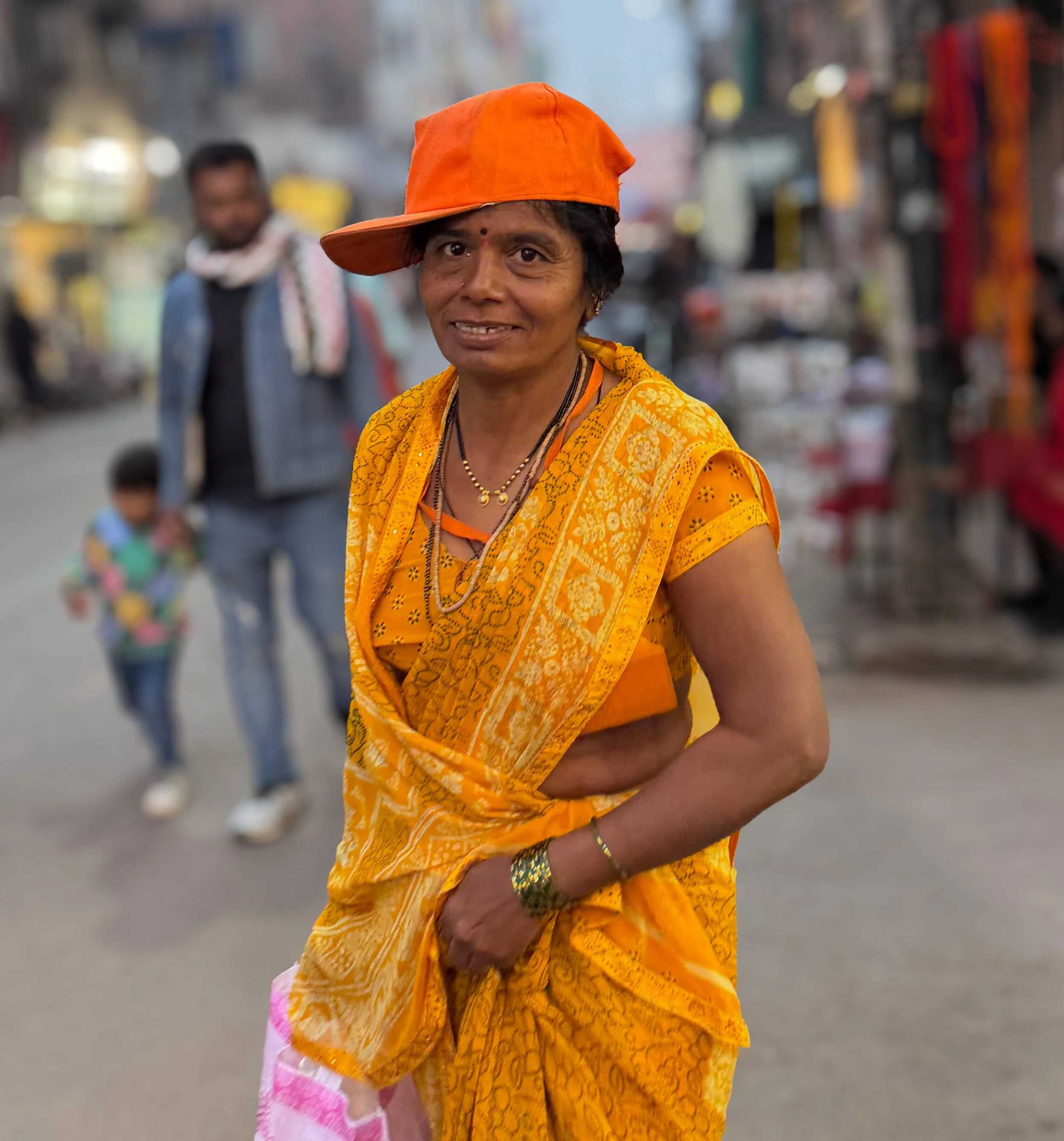A woman dressed in bright orange traditional Indian attire with an orange hat, standing on a busy street with blurred pedestrians and shops in the background.
