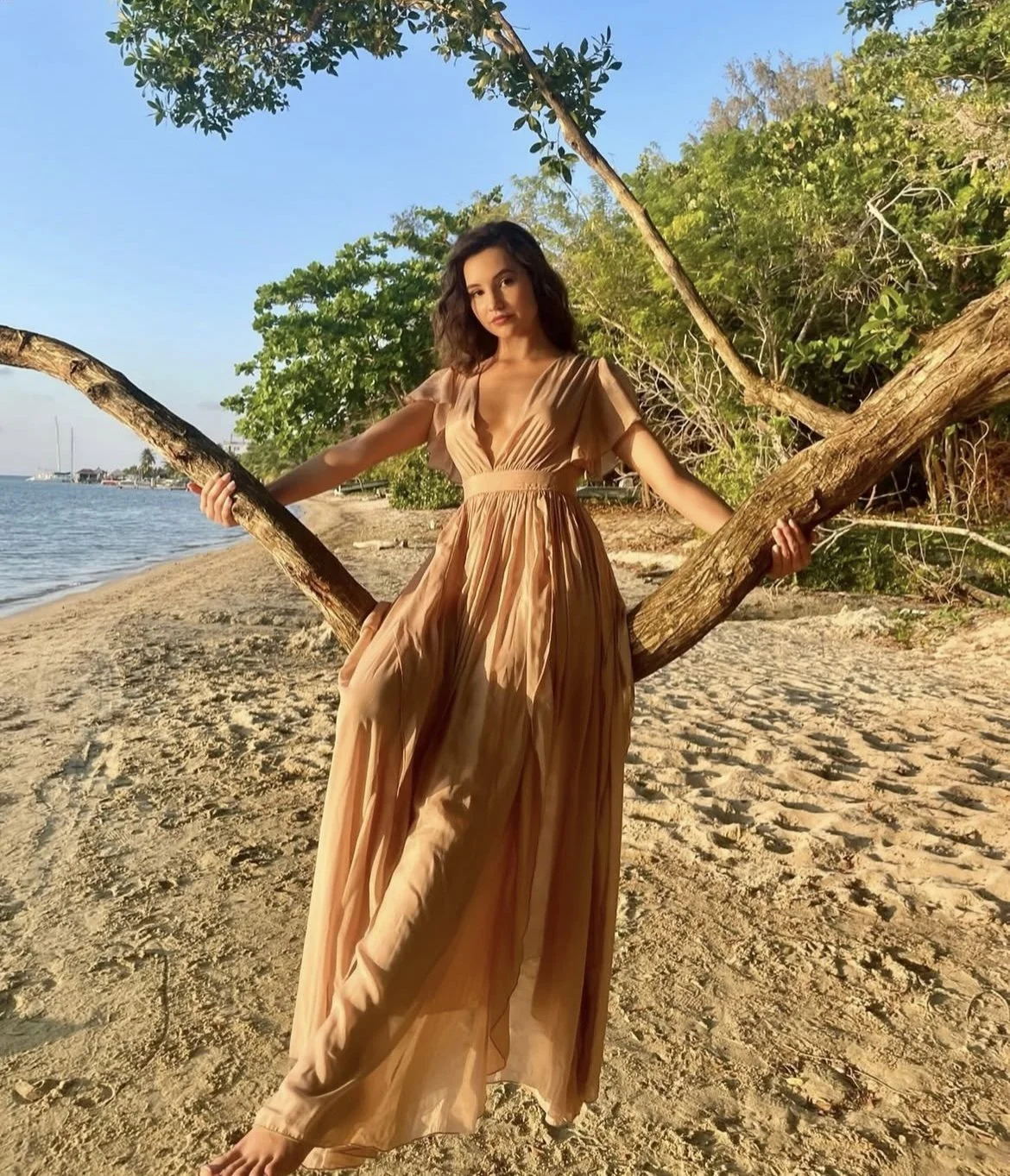A woman in a tan dress standing on a sandy beach holding onto a tree branch. There is water and trees in the background. Roatan
