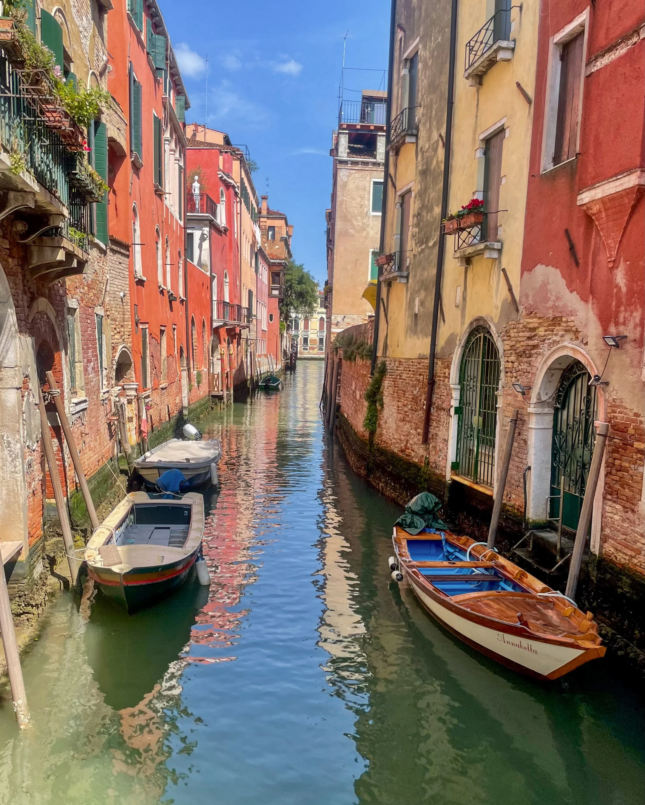 Colorful buildings lining a narrow canal with boats docked along the sides, clear blue sky overhead.