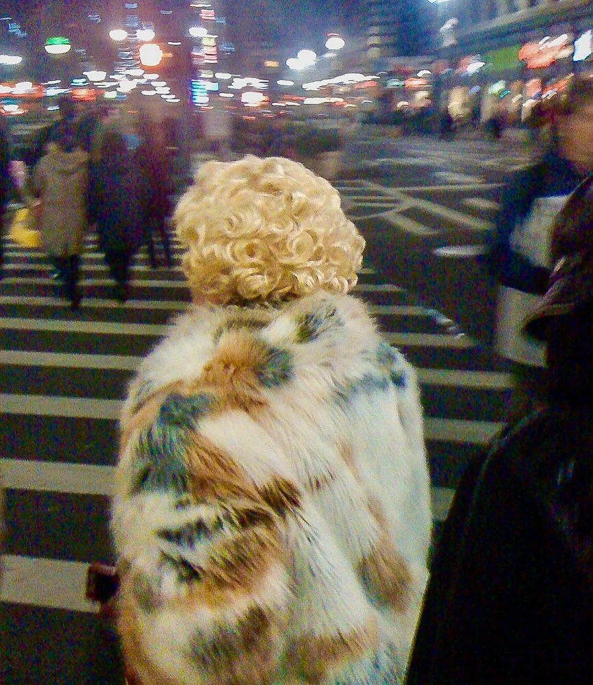 Person with curly blonde hair and a colorful furry coat standing at a busy crosswalk at night in an urban area.