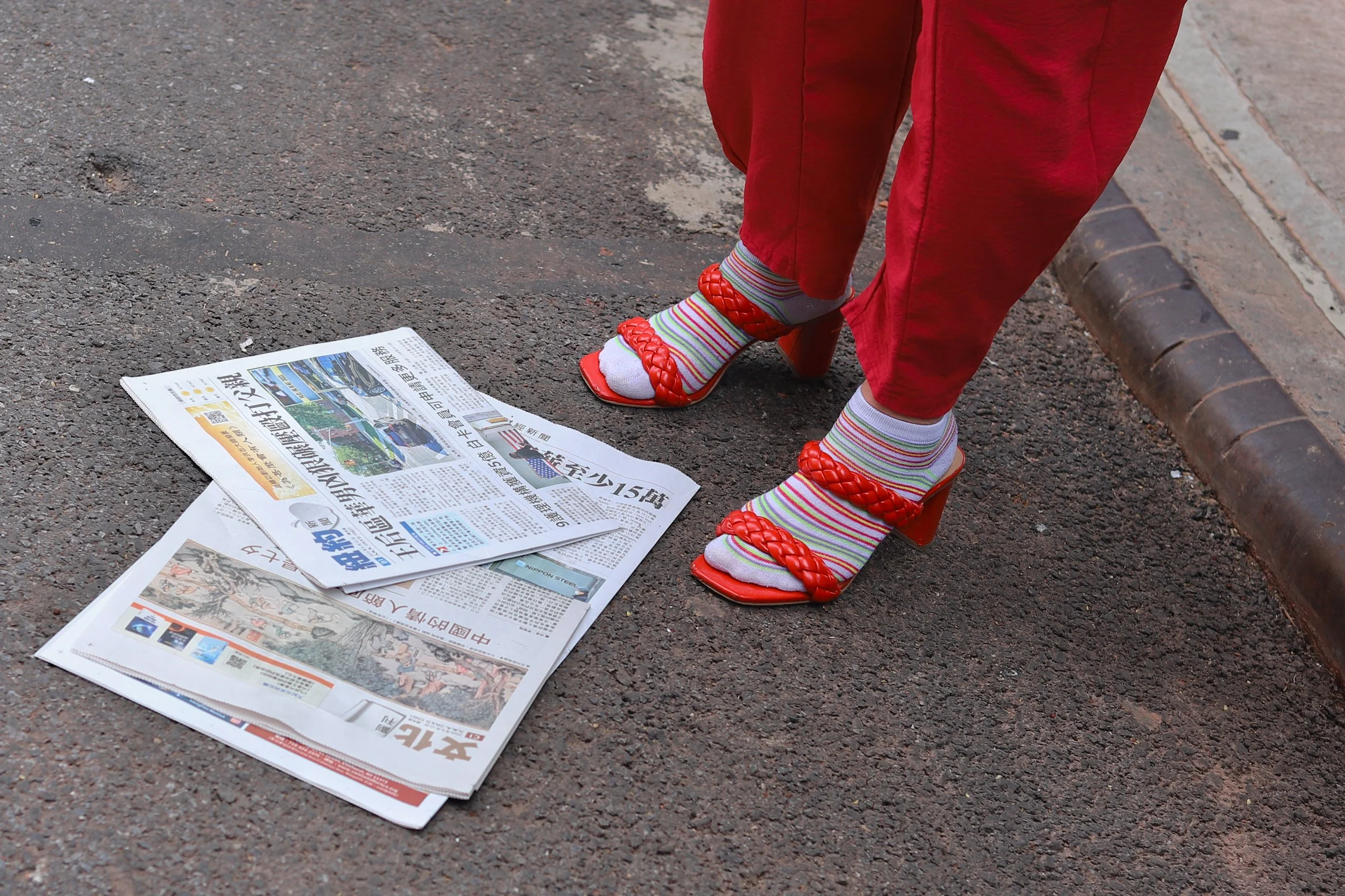 Close-up of a person's legs wearing red pants and colorful striped socks with red high-heeled sandals, standing over newspapers on a paved sidewalk.