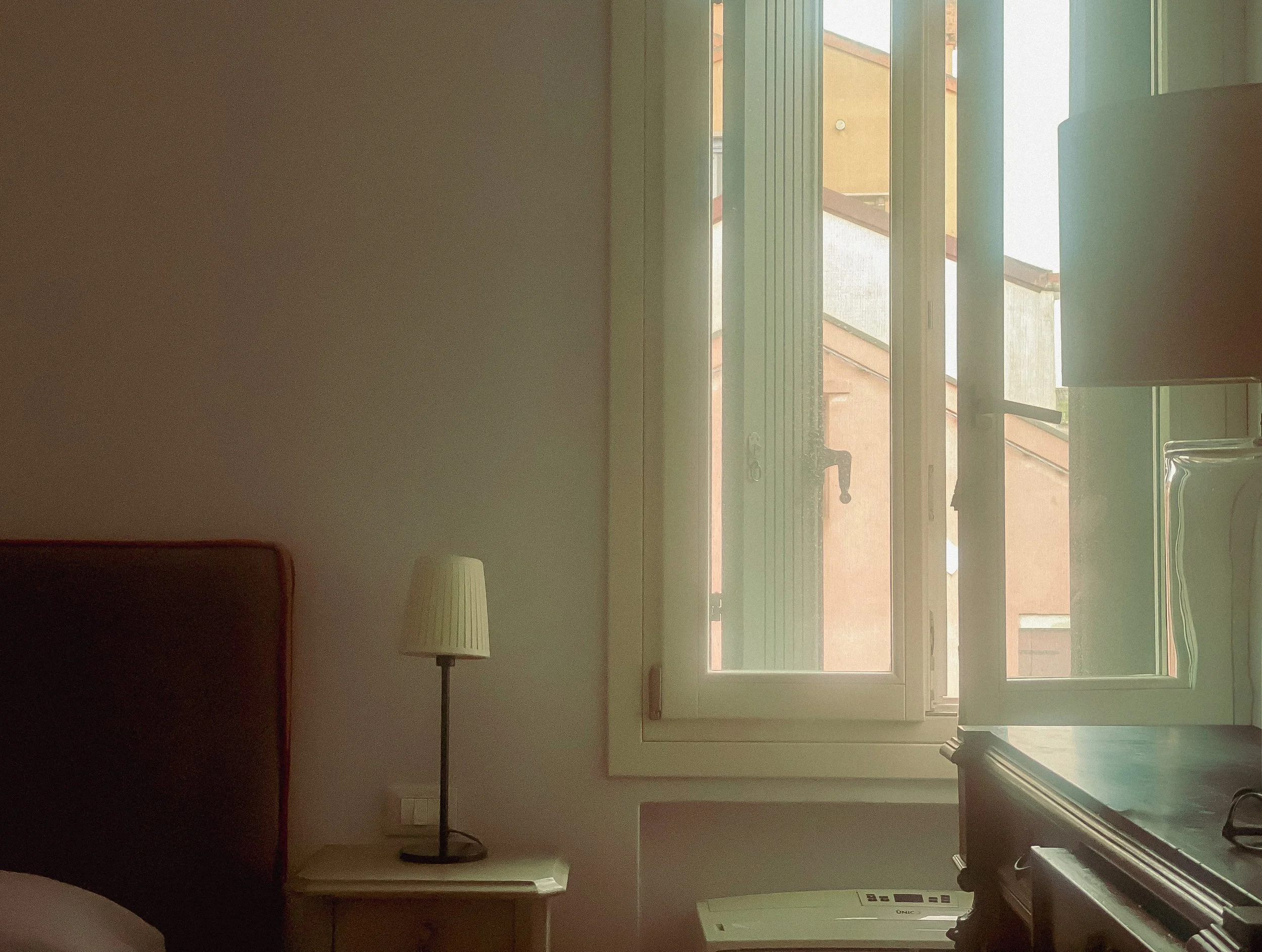 A hotel room with a window showing an outside view of neighboring buildings and stairs. Inside, there is a bed, a bedside table with a lamp, and a desk with a lamp and glasses.