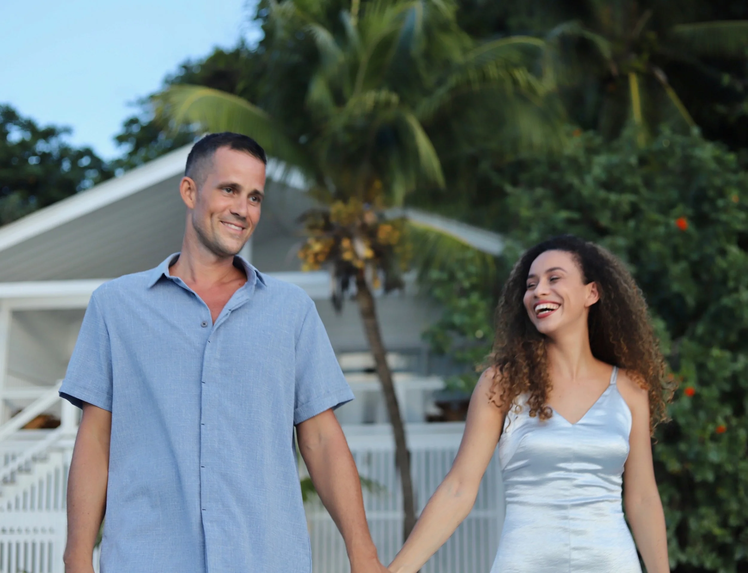 A young couple holding hands and smiling outdoors in a tropical setting with palm trees and a house in the background.