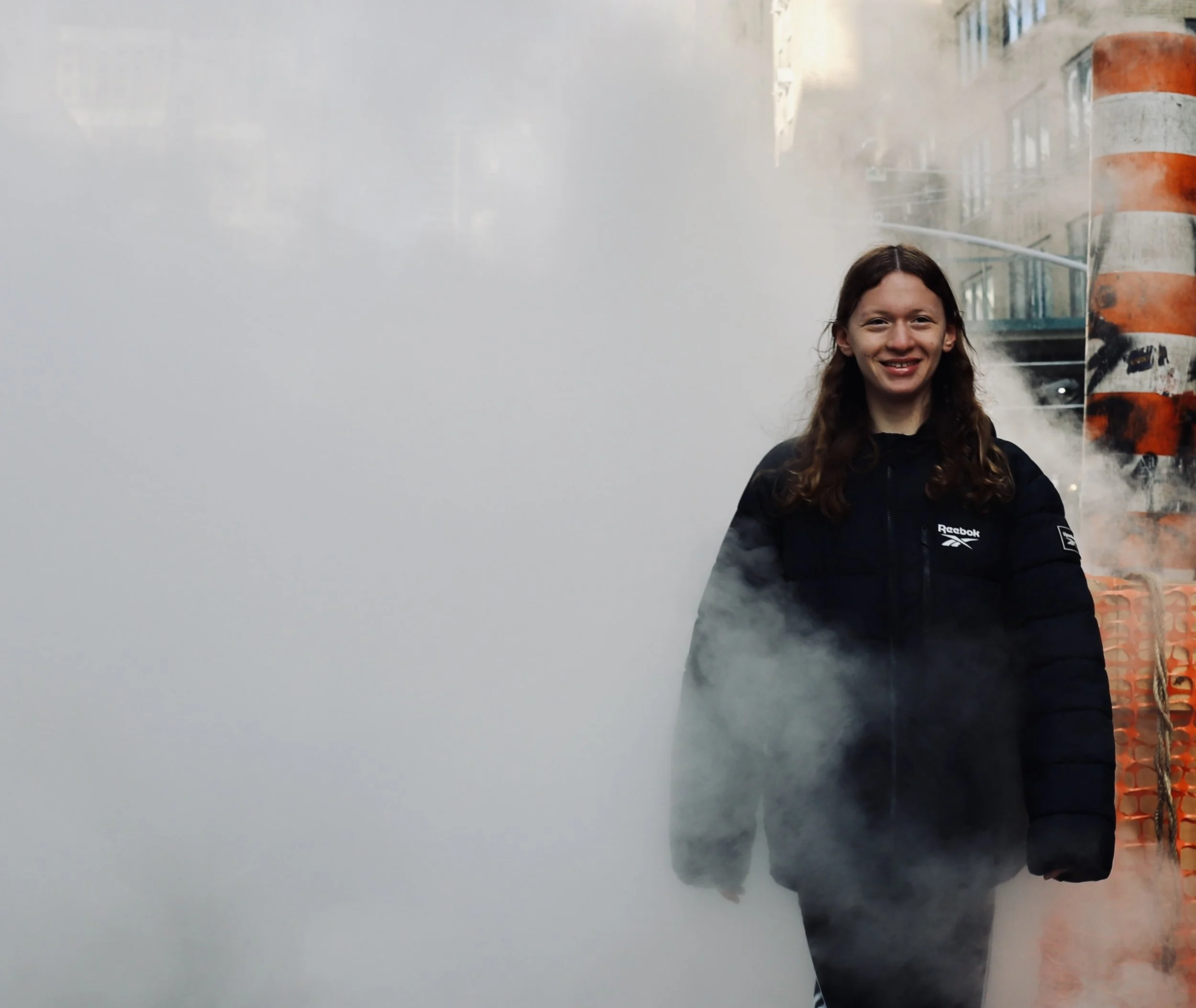 Smiling woman with long hair wearing a black Reebok jacket standing amidst white steam or fog outdoors, with orange construction barrels in the background.
