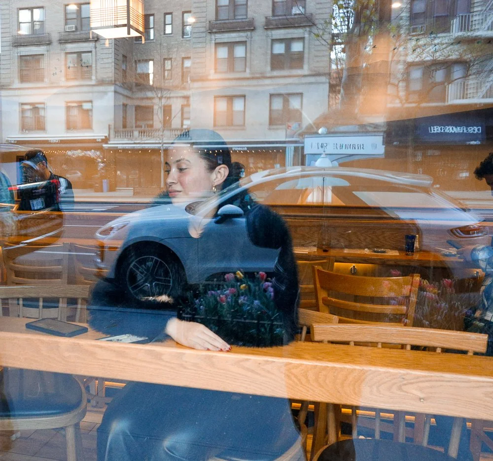 A woman sitting inside a cafe, seen through the window, with reflections of the street, cars, and buildings outside.
