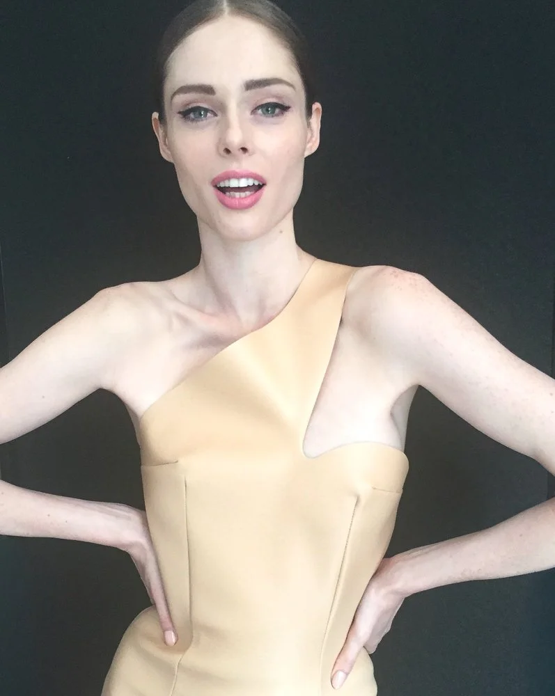 A young woman with fair skin, blue eyes, and brown hair styled back, wearing a beige one-shoulder dress, standing against a black background with her hands on her hips and smiling slightly.